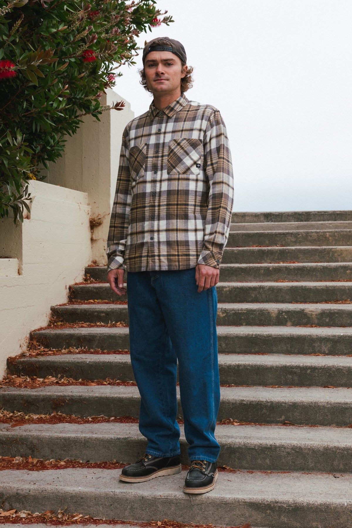 A man wearing a plaid button-down shirt, 686 Men’s Unwork® Hammerhead Denim Pant, and dark boots standing on concrete steps.