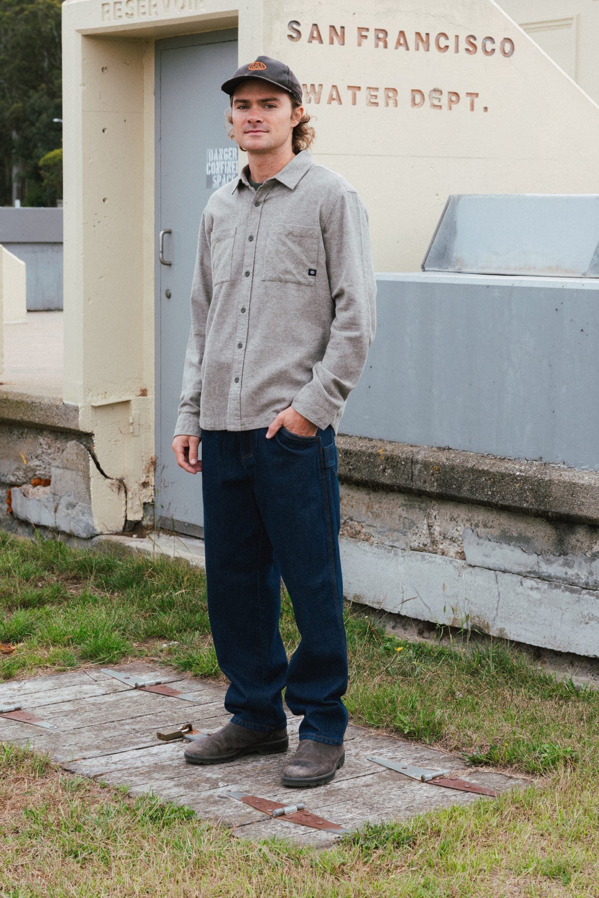 A man wearing a grey long-sleeved button-down shirt, 686 Men’s Unwork® Hammerhead Denim Pant, and a baseball cap standing outdoors.