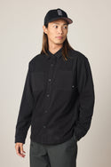A person with long brown hair wears the 686 Men's Whisper Knit Flannel and a black baseball cap, paired with dark pants, standing against a plain light background and looking to the side with one hand in their pocket.