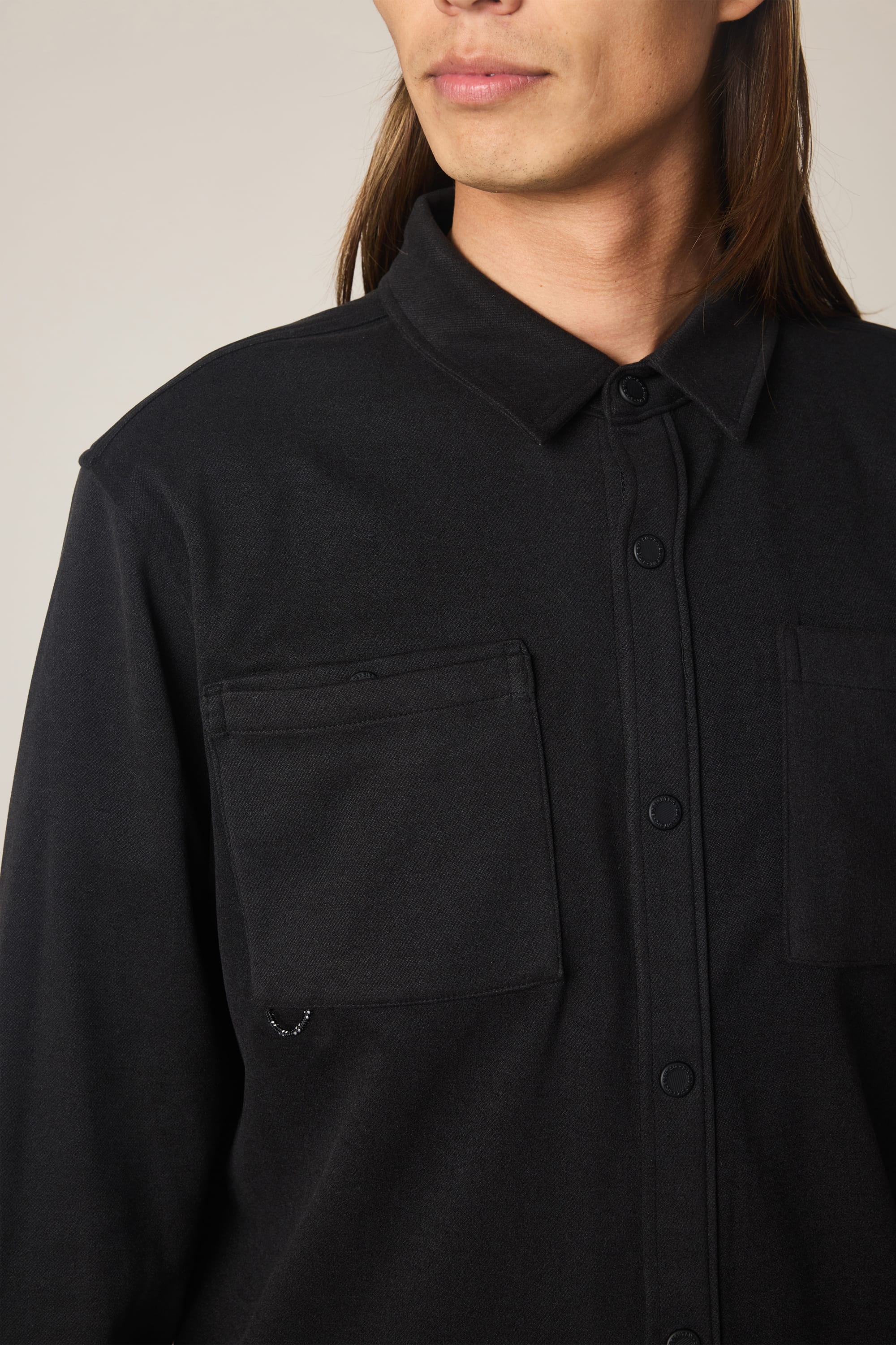 A person with long brown hair models the 686 Men's Whisper Knit Flannel by 686, a black polyester shirt with two breast pockets. Only the lower half of their face and upper torso are visible against a plain light background.