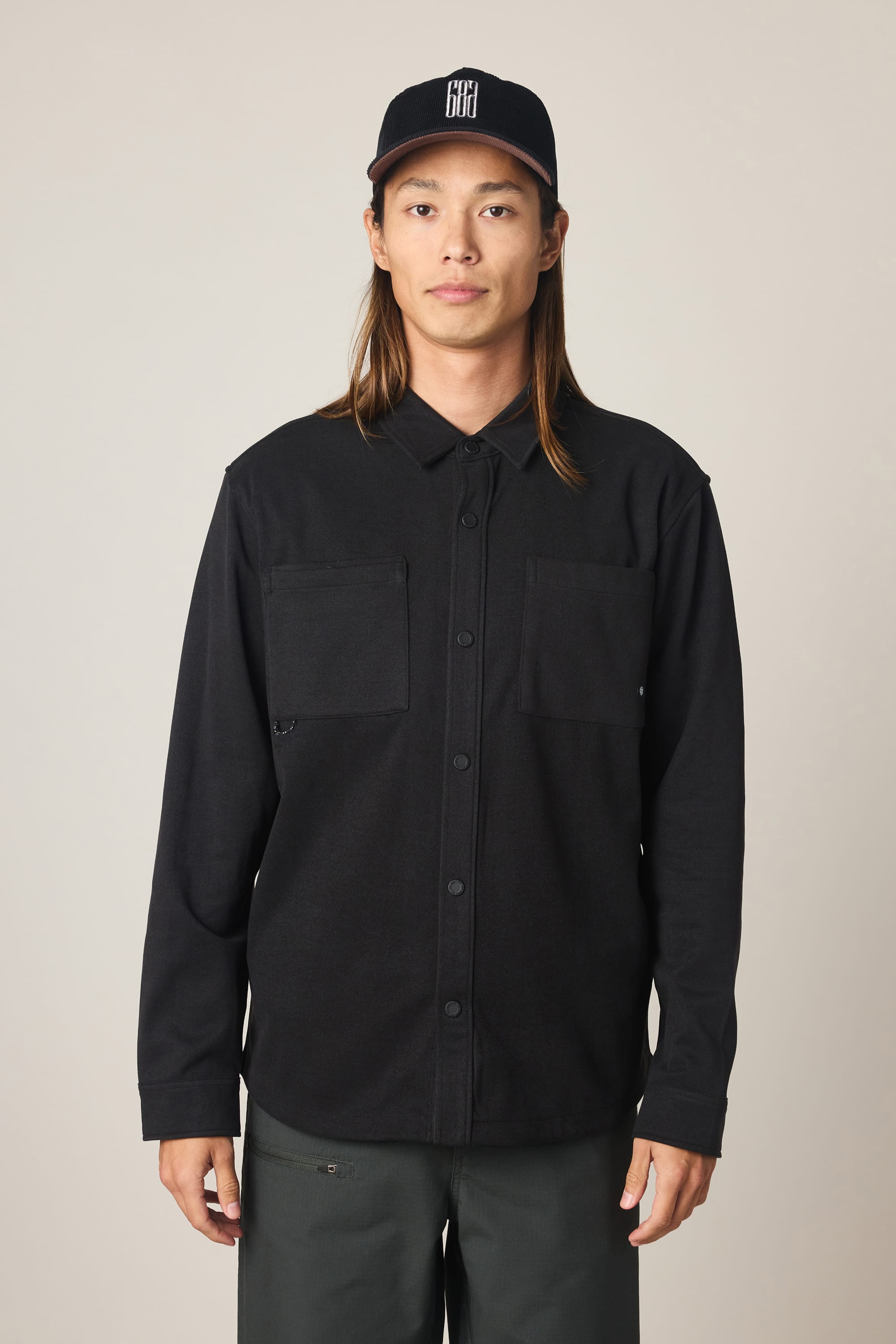 A person with long hair is standing facing forward, wearing the 686 Men's Whisper Knit Flannel by 686, paired with dark pants and a black baseball cap featuring a white logo. The background is plain and light-colored.