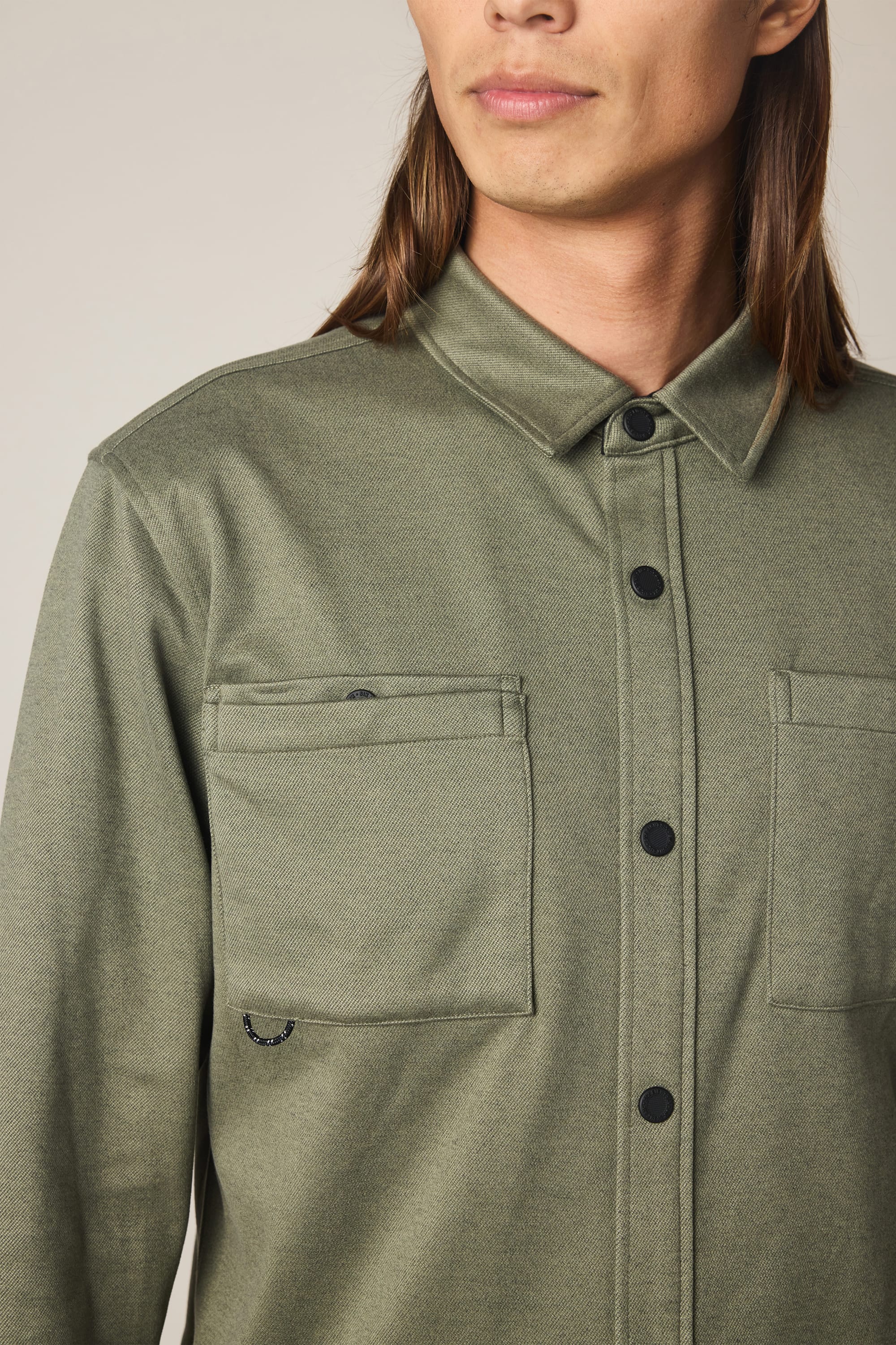 A person with long hair wears the 686 Men's Whisper Knit™ Flannel in olive green, a button-up shirt featuring two front pockets by 686, facing slightly to the right and shown from mid-chest up.