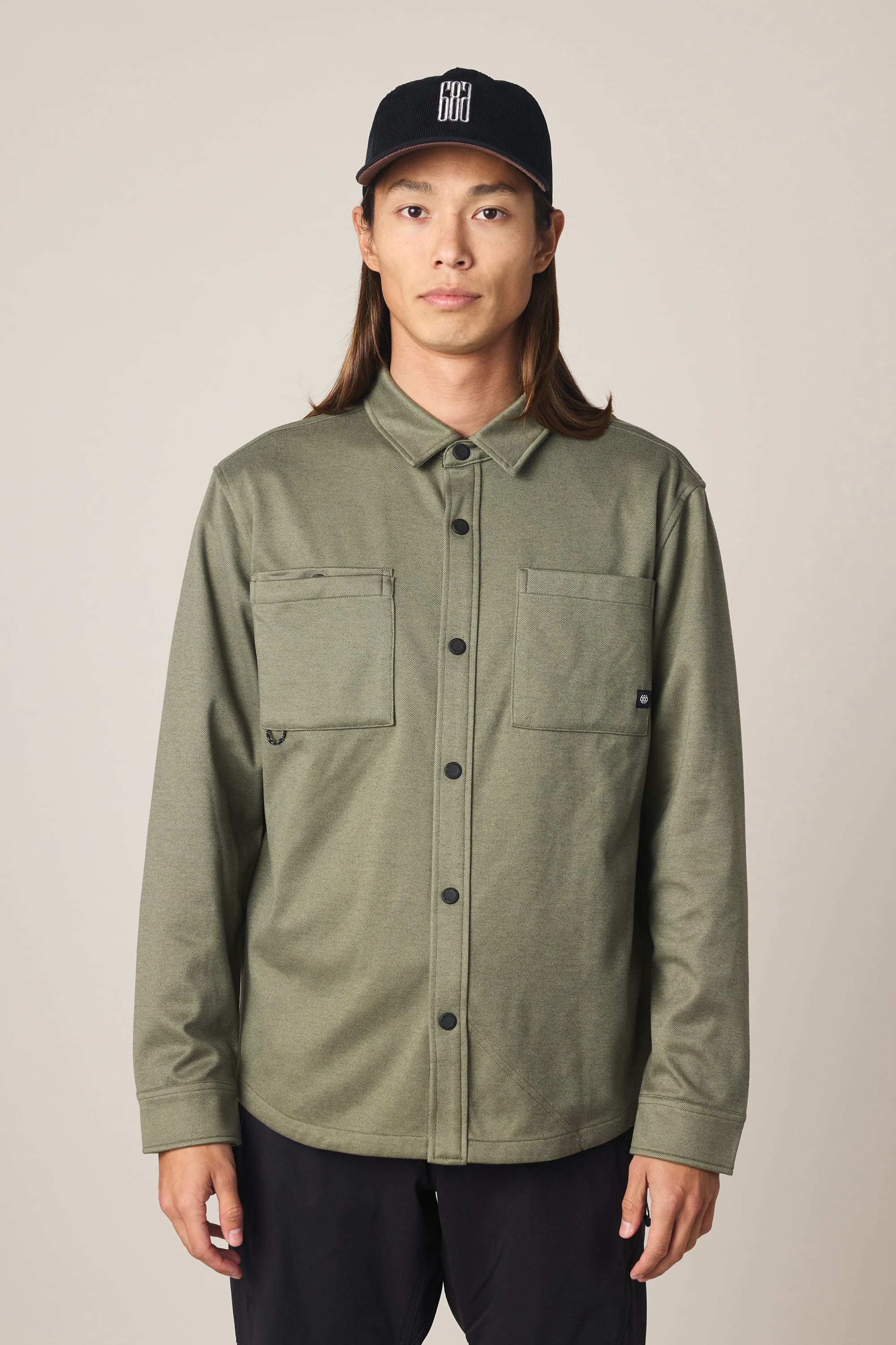 A person with long hair wearing a black cap, dressed in the 686 Men's Whisper Knit™ Flannel by 686—an olive green men's shirt crafted from 100% polyester with two front pockets—and black pants stands facing the camera against a plain background.