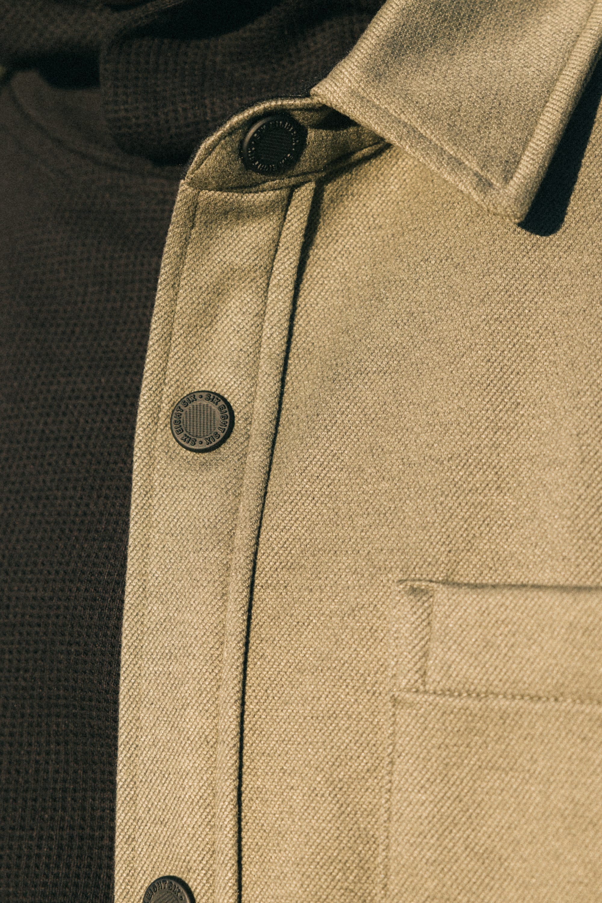 Close-up of the 686 Men's Whisper Knit™ Flannel in beige, featuring metal buttons and a chest pocket, worn over a dark ribbed men's shirt.