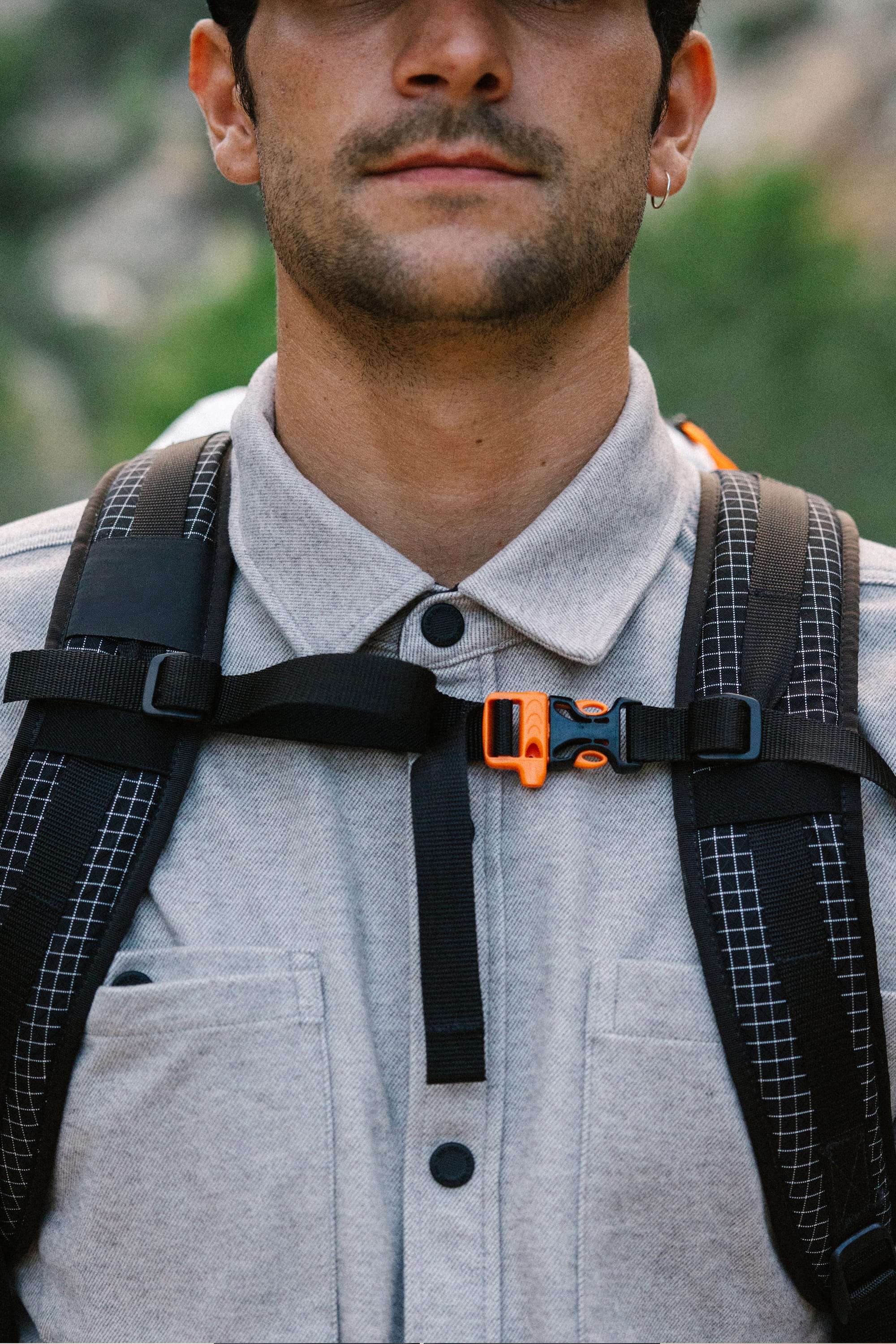A person wearing the 686 Men's Whisper Knit™ Flannel shirt, featuring button-up styling and black backpack straps with an orange buckle, stands outdoors with blurred greenery in the background.