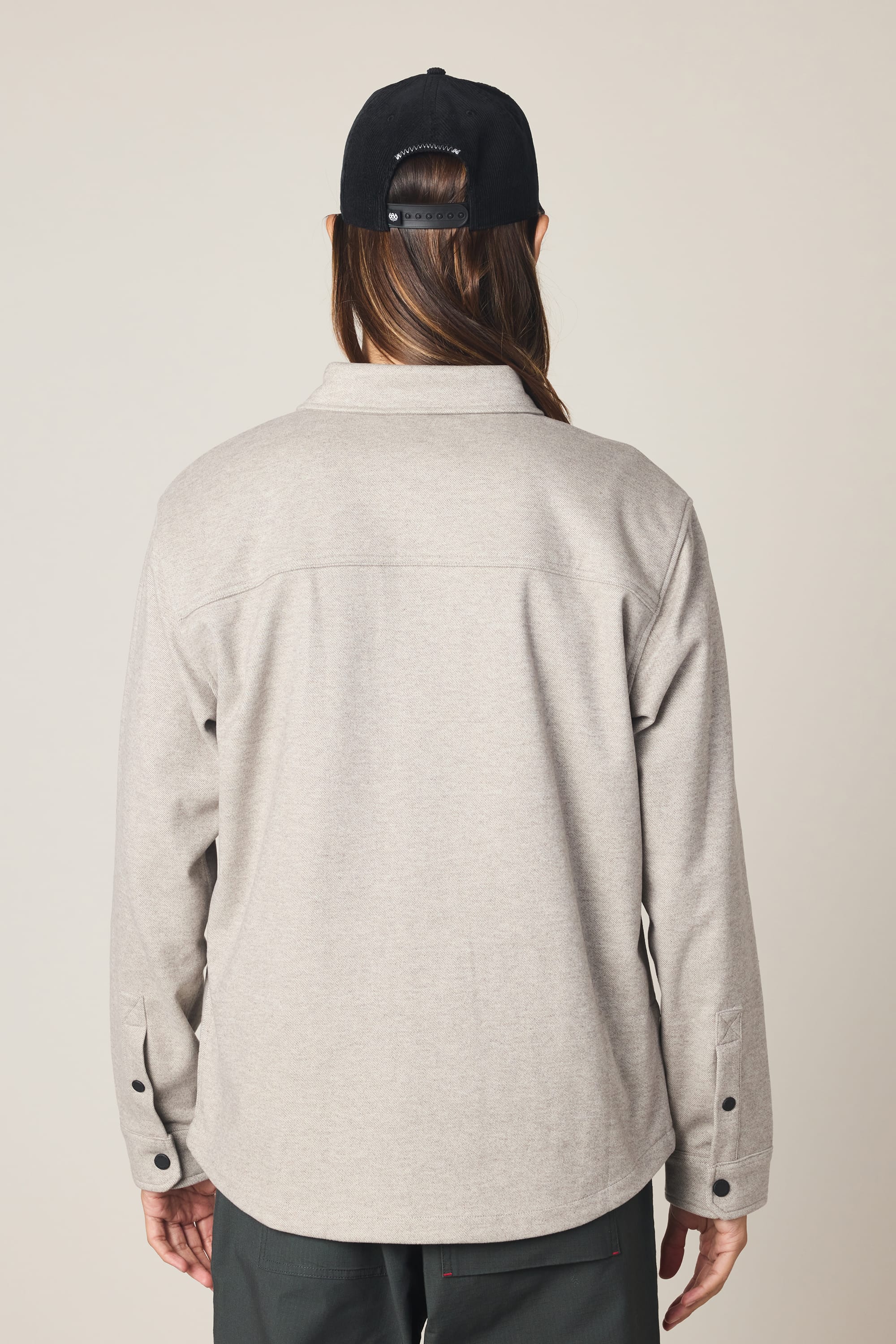 A person with long hair, wearing a black baseball cap, dark pants, and the 686 Men's Whisper Knit Flannel by 686 in light gray, is seen from behind against a plain light background.