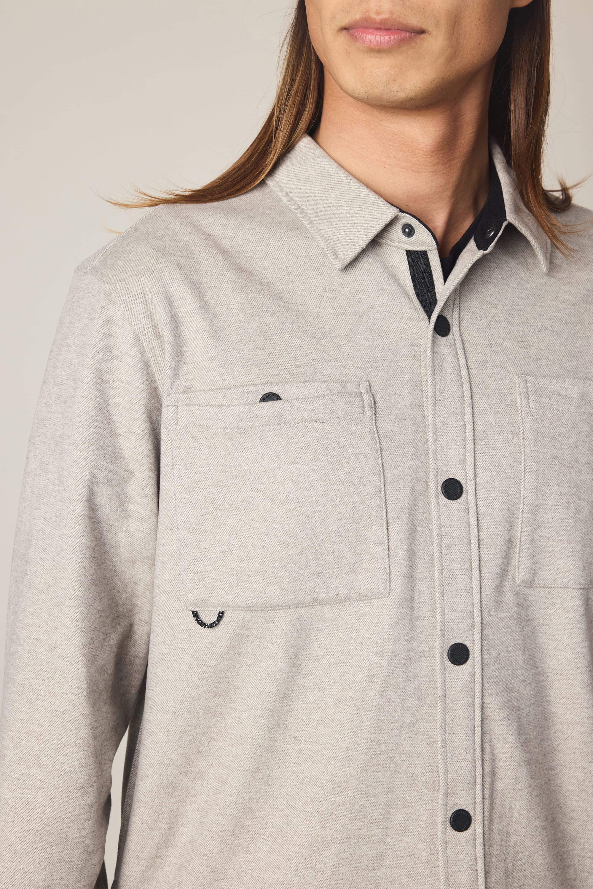 A person with long hair wears the 686 Men's Whisper Knit Flannel in light gray, featuring two chest pockets. Standing against a plain background, their face is partially visible as they look to the side.
