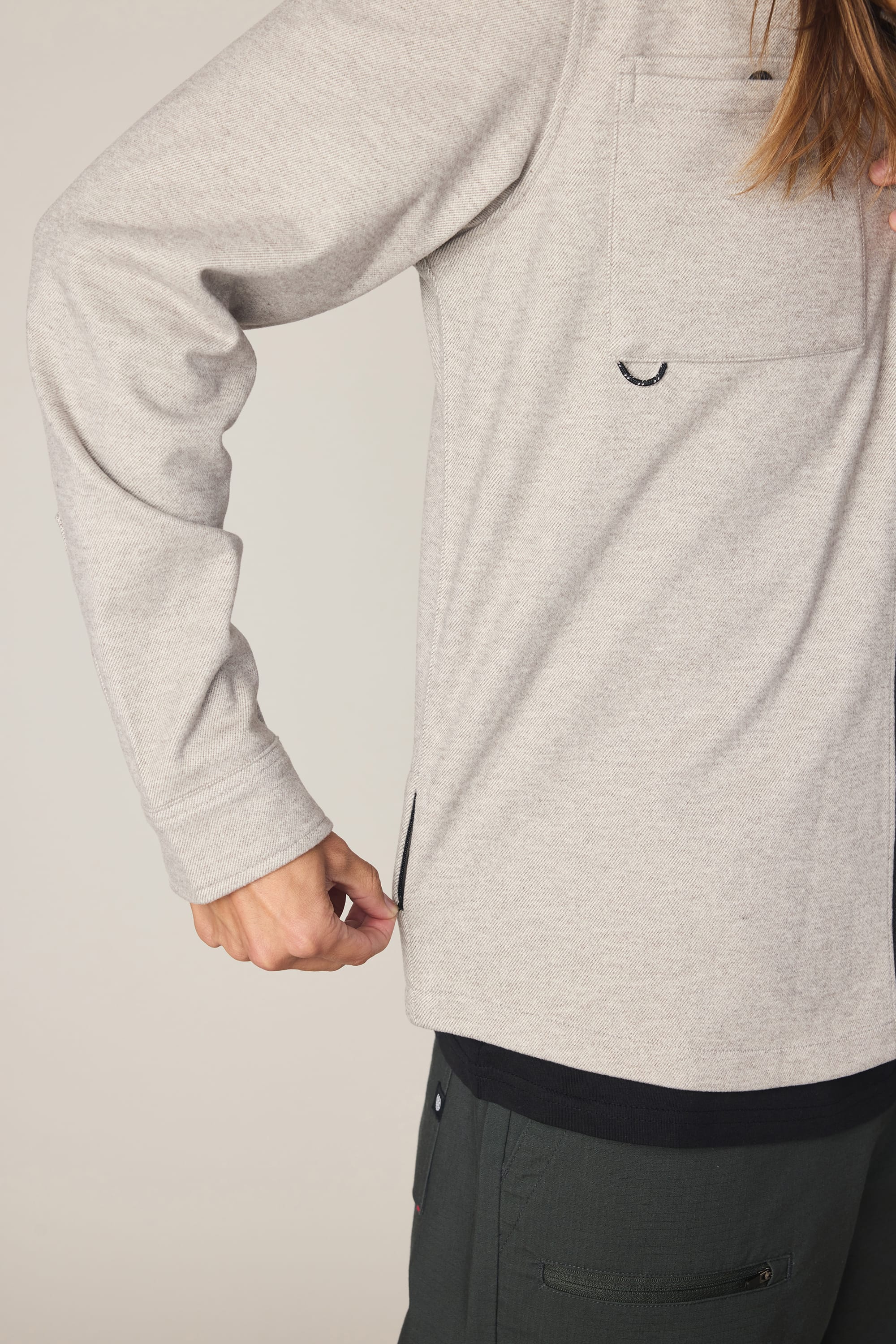 A person wearing a 686 Men's Whisper Knit Flannel in light gray and dark pants pulls on a small side seam zipper near the hem, with only their torso and arm visible.