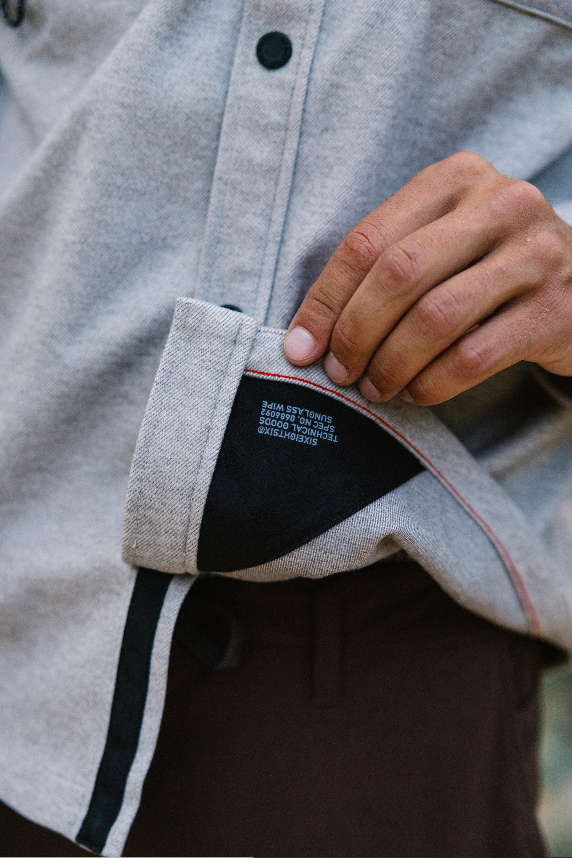 A person lifts the corner of a light gray 686 Men's Whisper Knit™ Flannel, revealing a black inner lining with white text. The shirt features black buttons and red stitching along the edge.