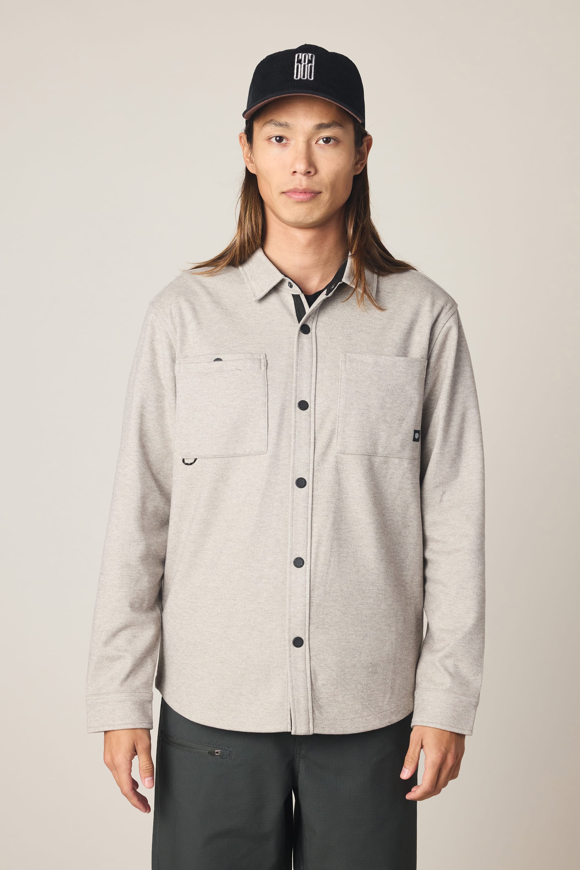 A person with long brown hair wears a black cap, dark pants, and a light gray 686 Men's Whisper Knit Flannel by 686, featuring two chest pockets, standing against a plain light background.