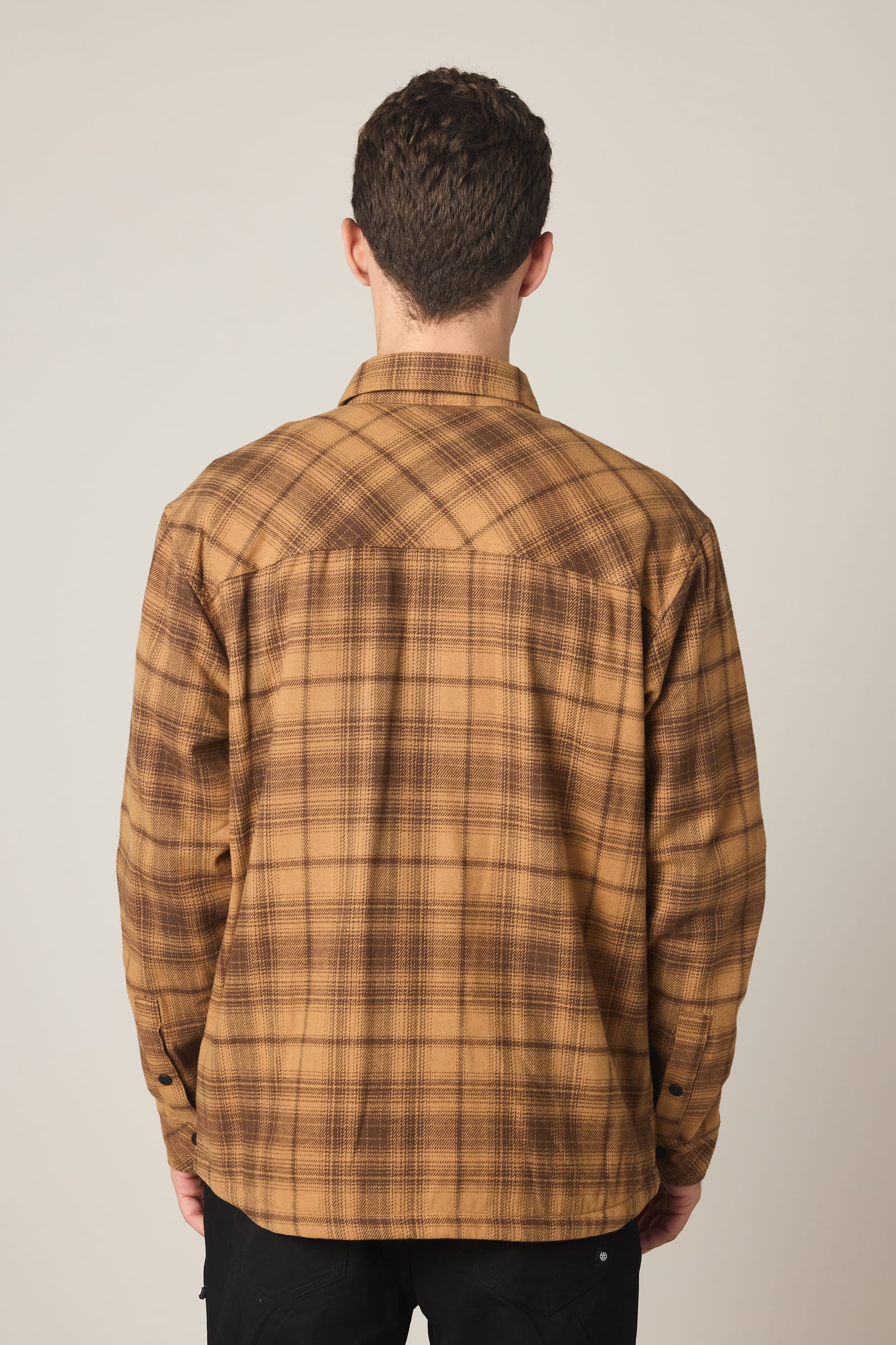 A person with short curly hair, seen from behind, wears a 686 Men's Heat Grid Flannel by 686 in brown and black plaid with black pants, standing against a plain light background.