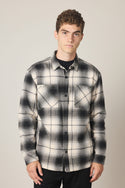A young man with short curly hair wears the 686 Men's Heat Grid Flannel by 686, a black and white plaid polyester shirt, standing against a plain light background and looking at the camera with a neutral expression.