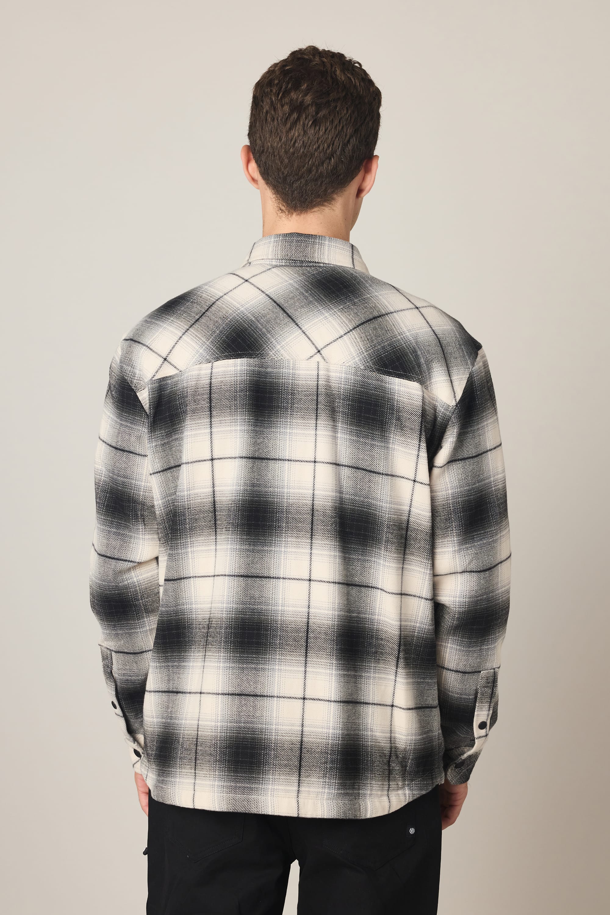 A person with short brown hair stands with their back to the camera, wearing a black and white 686 Men's Heat Grid Flannel shirt and black pants against a plain light background.