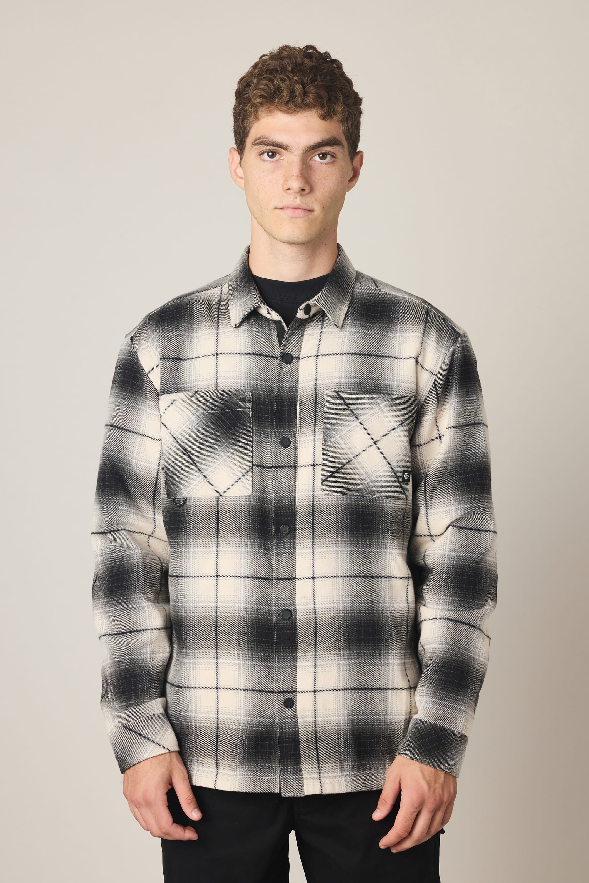 A person stands facing forward, wearing the 686 Men's Heat Grid™ Flannel by 686—a black-and-white plaid button-up shirt made from 100% polyester. It is layered over a black top, with the wearer displaying a neutral expression against a light background.