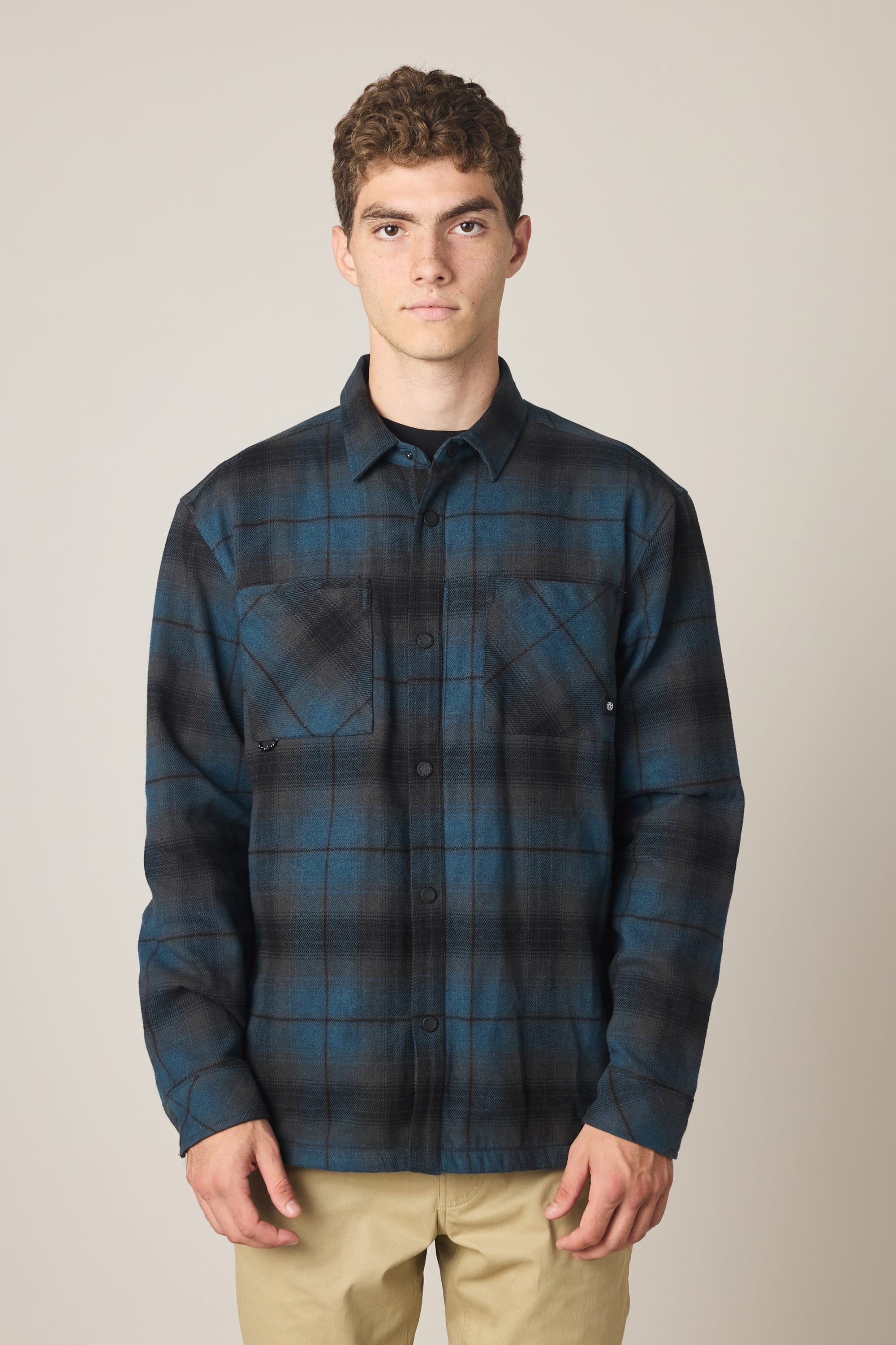 A young man with short curly hair is wearing the 686 Men's Heat Grid™ Flannel shirt by 686 in a blue and black plaid pattern, paired with beige pants. He stands against a plain light background, looking directly at the camera with a neutral expression.