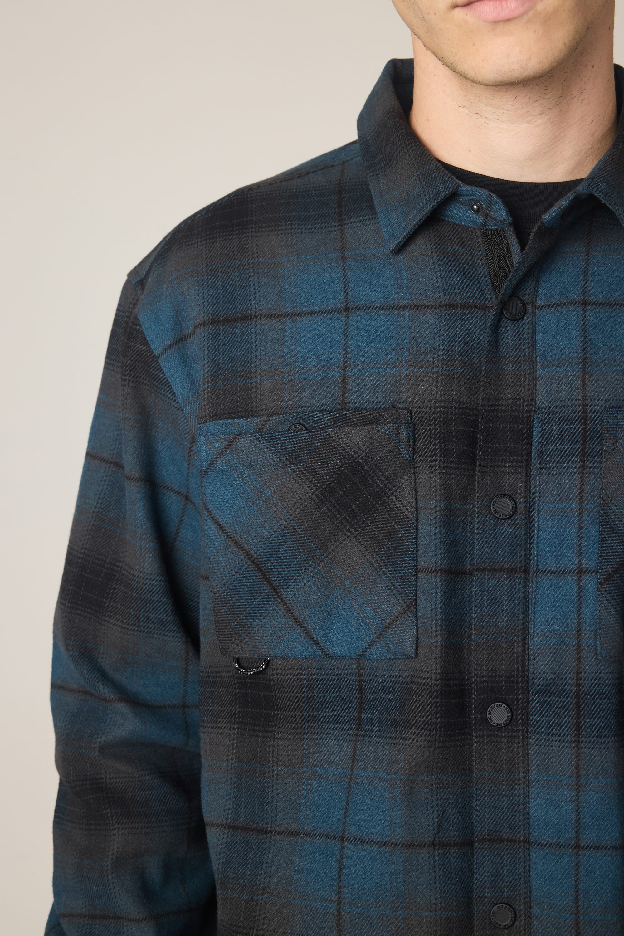 Close-up of a person wearing the 686 Men's Heat Grid Flannel in blue and black polyester with a chest pocket. The face is partially out of frame, set against a neutral background.