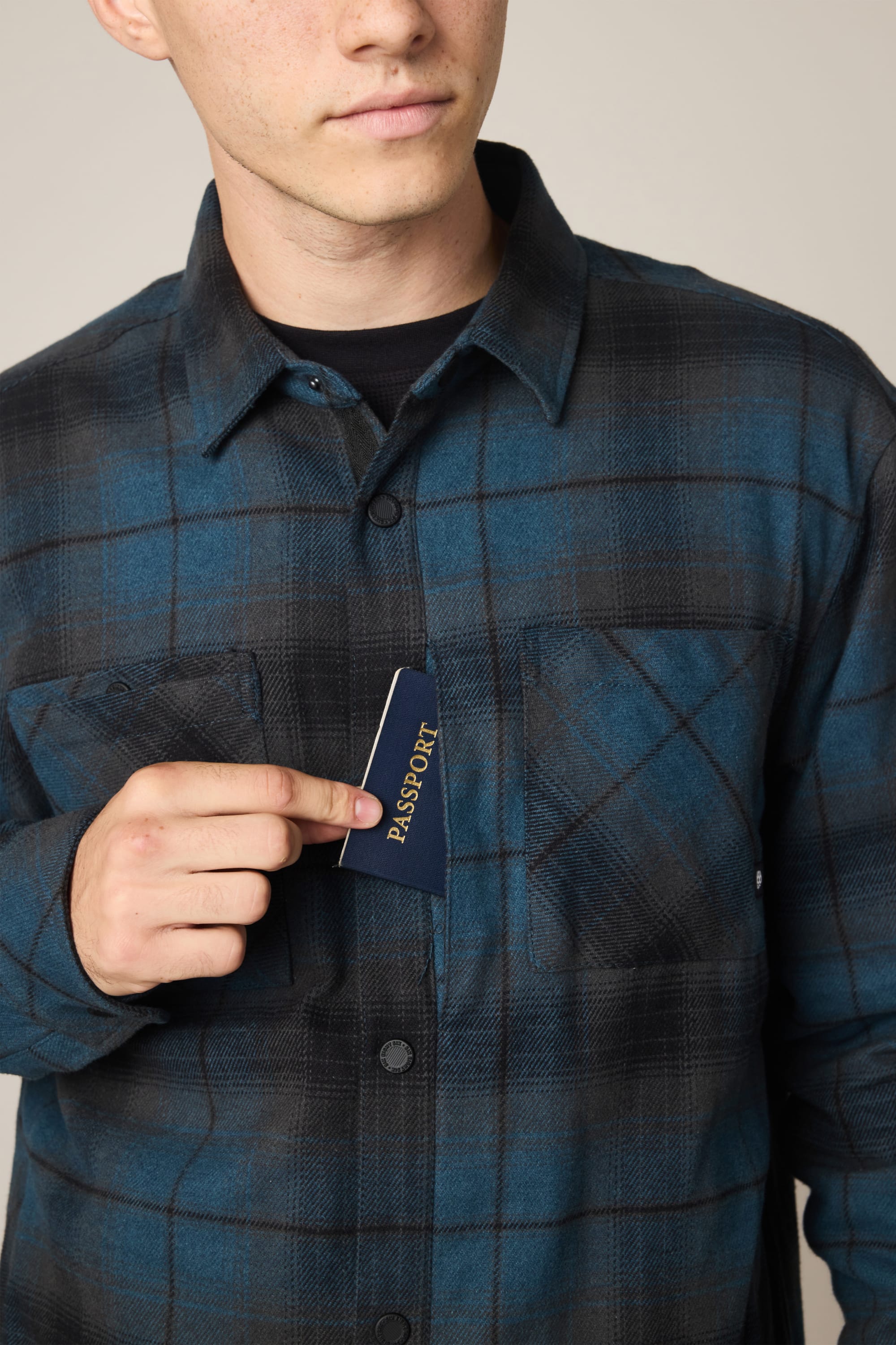 A person wearing the 686 Men's Heat Grid™ Flannel in blue plaid slips a passport into its chest pocket, with only the lower half of their face visible.