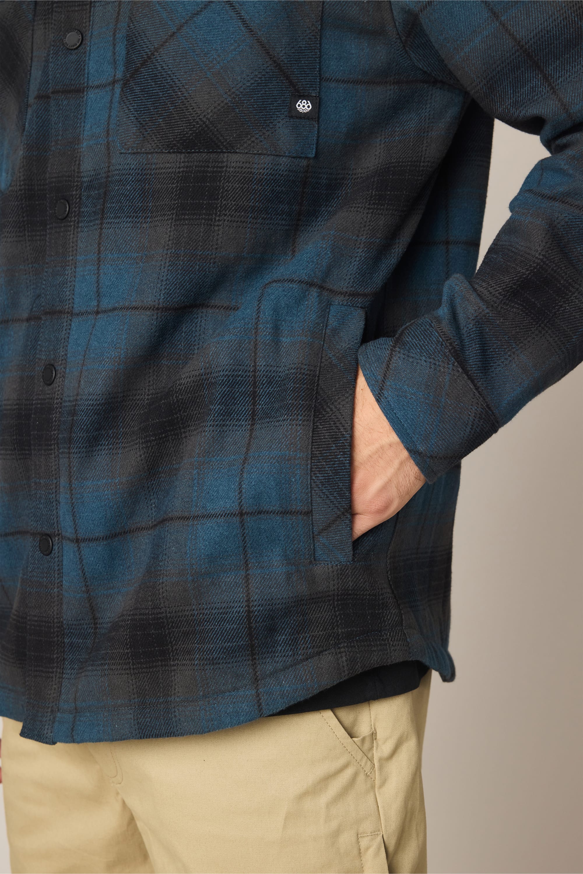 A person wearing the 686 Men's Heat Grid™ Flannel in a blue and black plaid pattern with one hand in the pocket, paired with beige pants. The photo is cropped to show only the torso and upper legs.