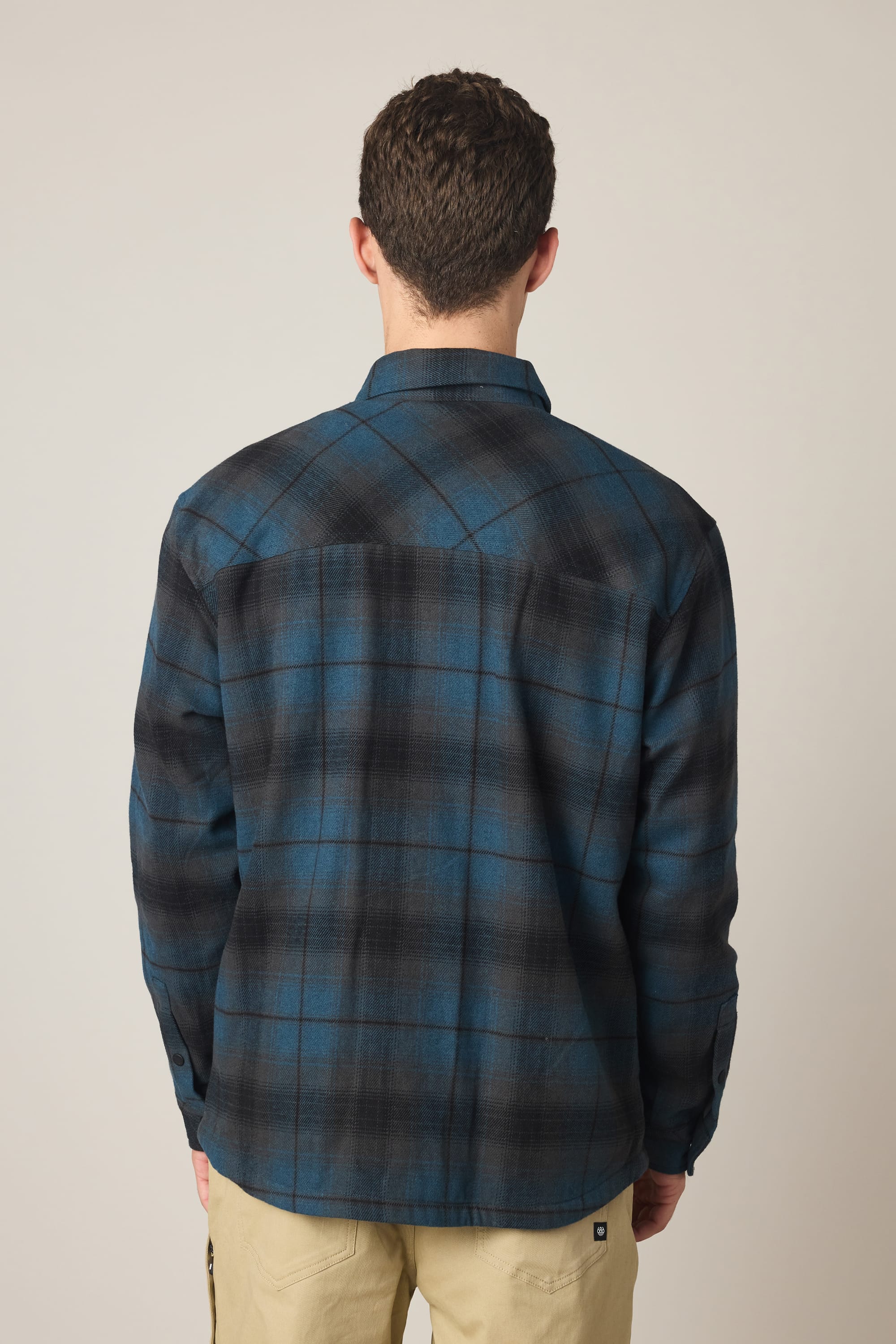 A man stands with his back to the camera, wearing the 686 Men's Heat Grid Flannel in blue and black plaid by 686, paired with khaki pants against a plain light background.