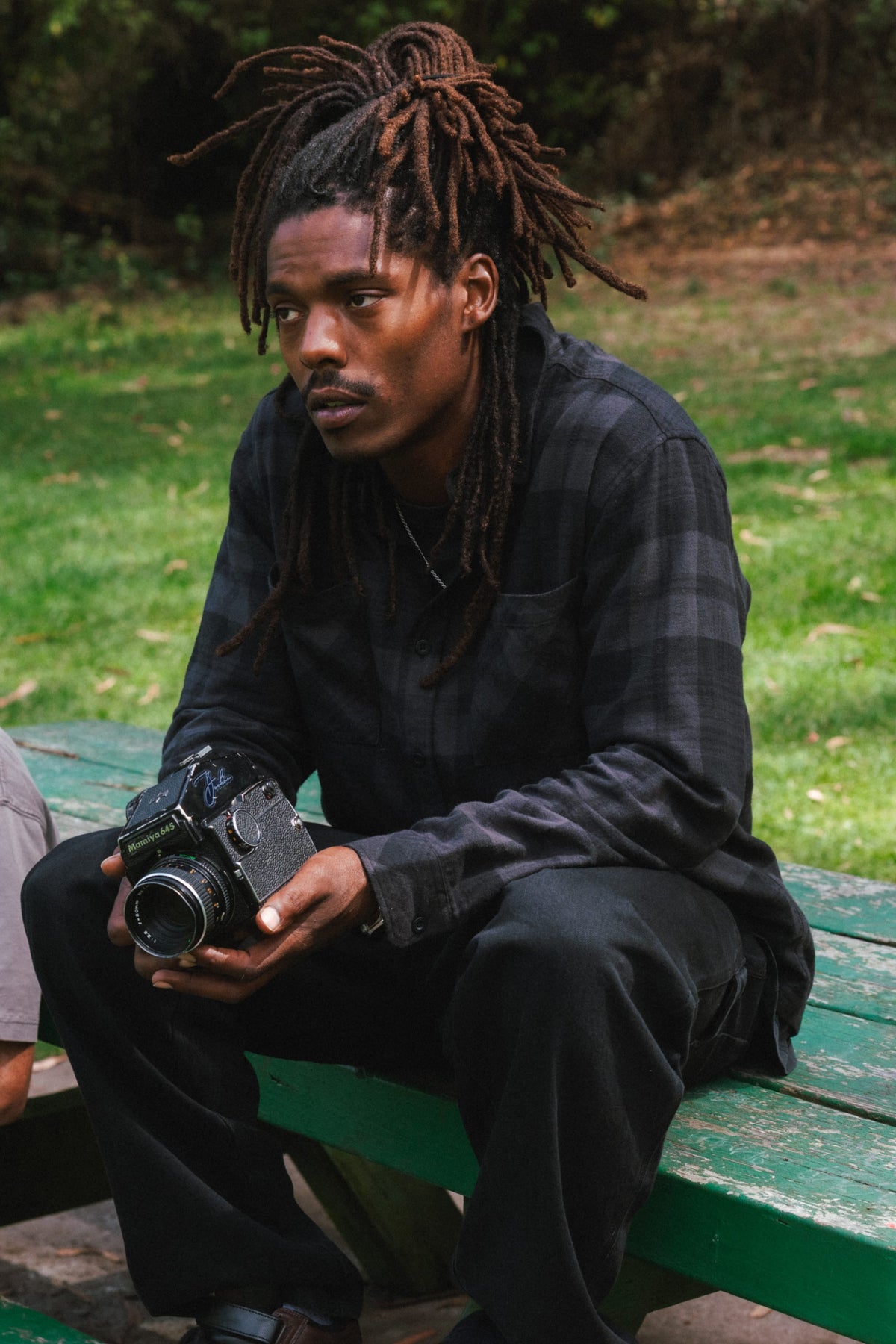 A man with dreadlocks wearing a dark grey 686 Men’s SMARTY® Headwall Flannel sits on a wooden bench holding a vintage film camera.