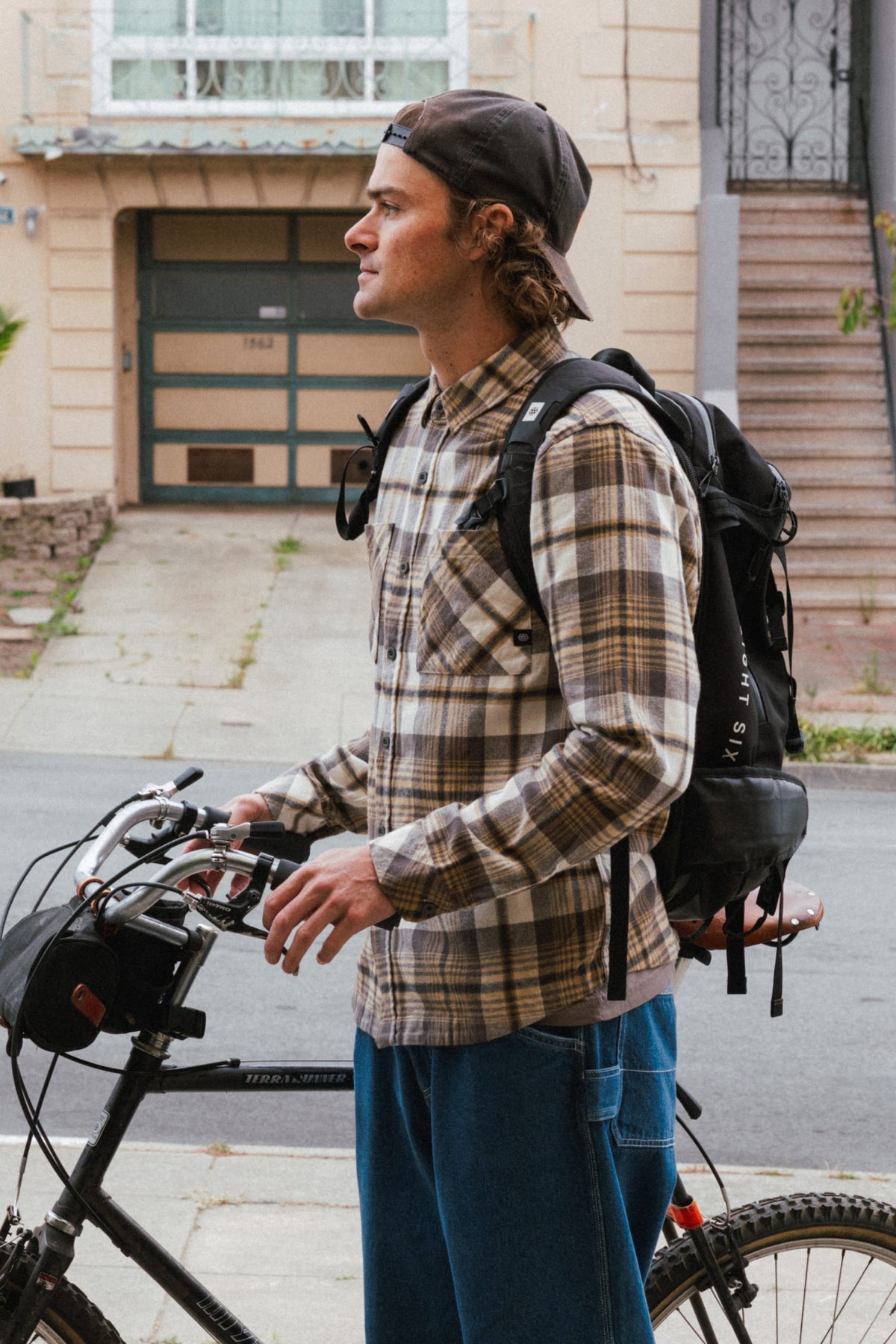 A man in a tan and brown 686 Men’s SMARTY® Headwall Flannel and a baseball cap stands holding the handlebars of a bicycle while wearing a backpack.