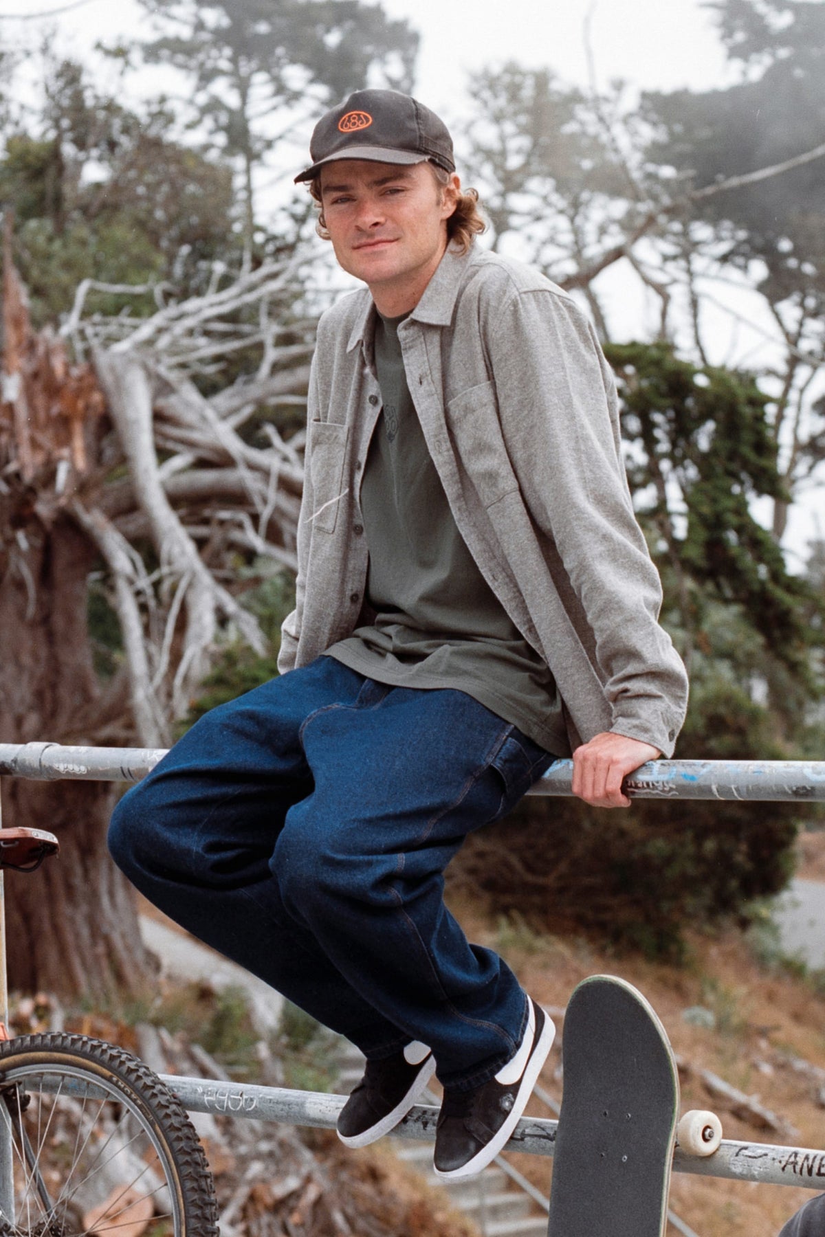 A man in a cap, 686 Men’s SMARTY® Headwall Flannel, and jeans sits on a metal railing next to a skateboard.