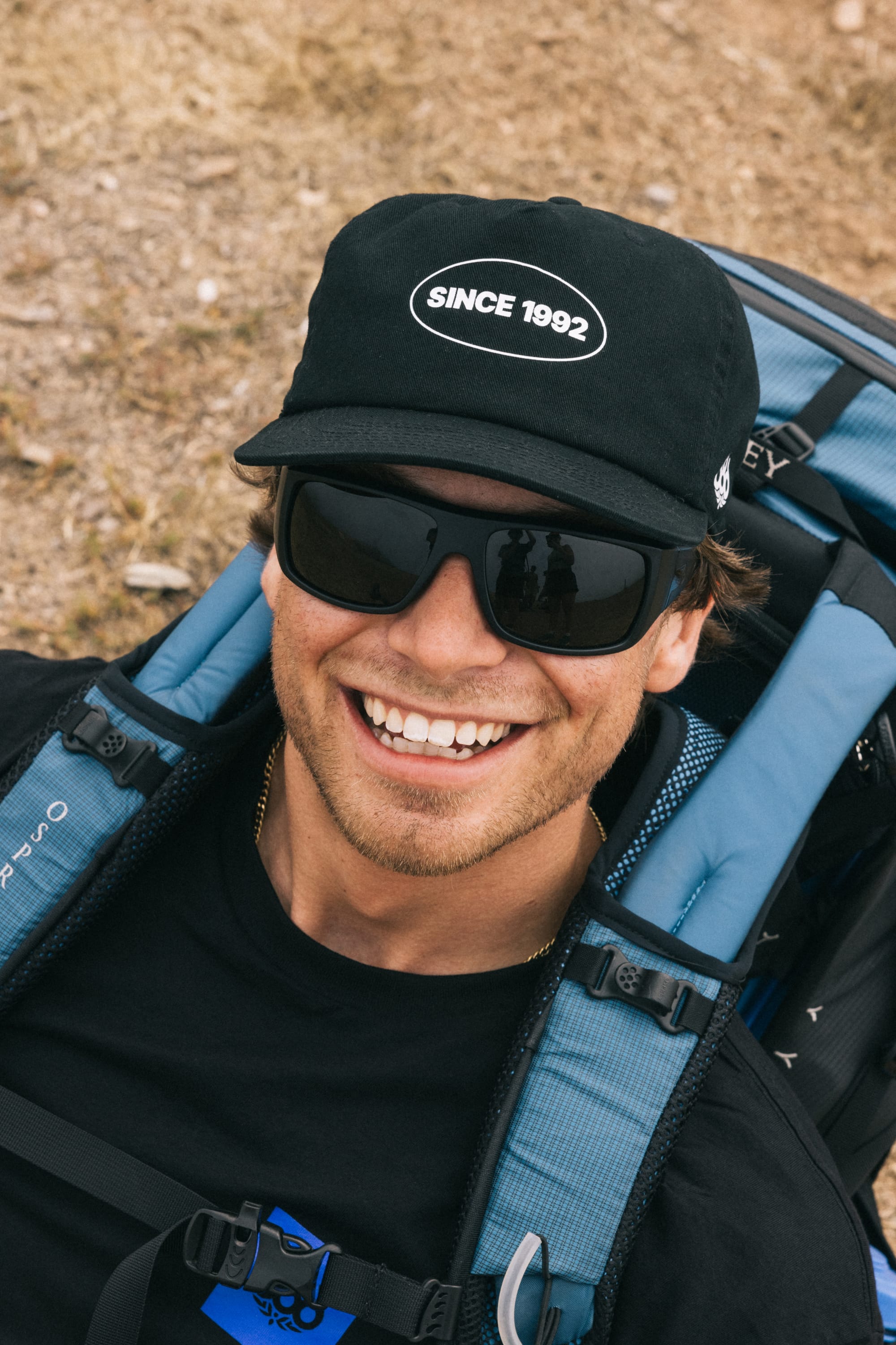 A smiling person wearing a black 100% cotton 686 1992 Unstructured Hat by 686, black sunglasses, and a blue backpack, outdoors on grass.