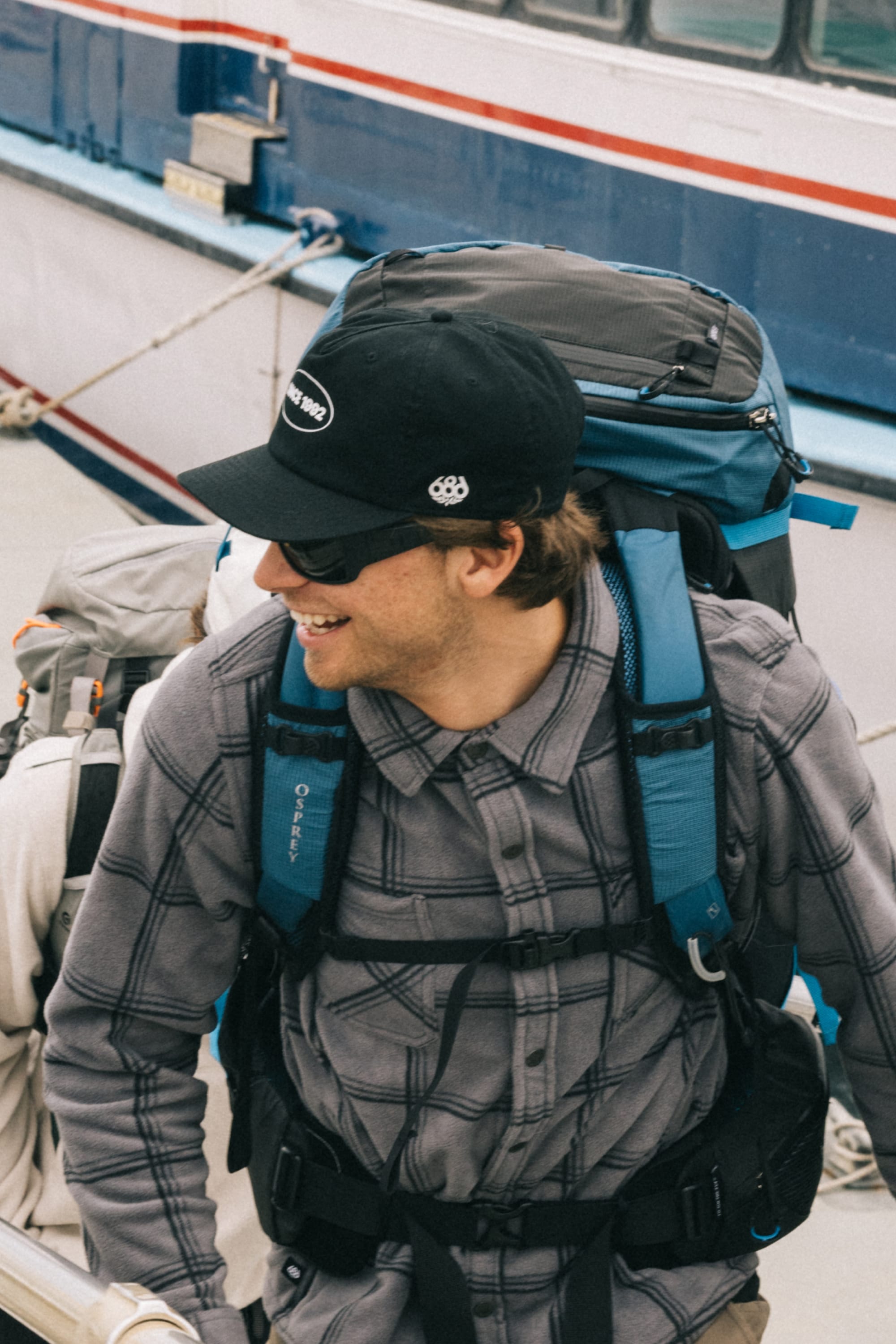 A smiling person wearing sunglasses, a gray plaid shirt, and the 686 1992 Unstructured Hat by 686, with a large blue backpack, stands near a boat painted white and blue with a red stripe.