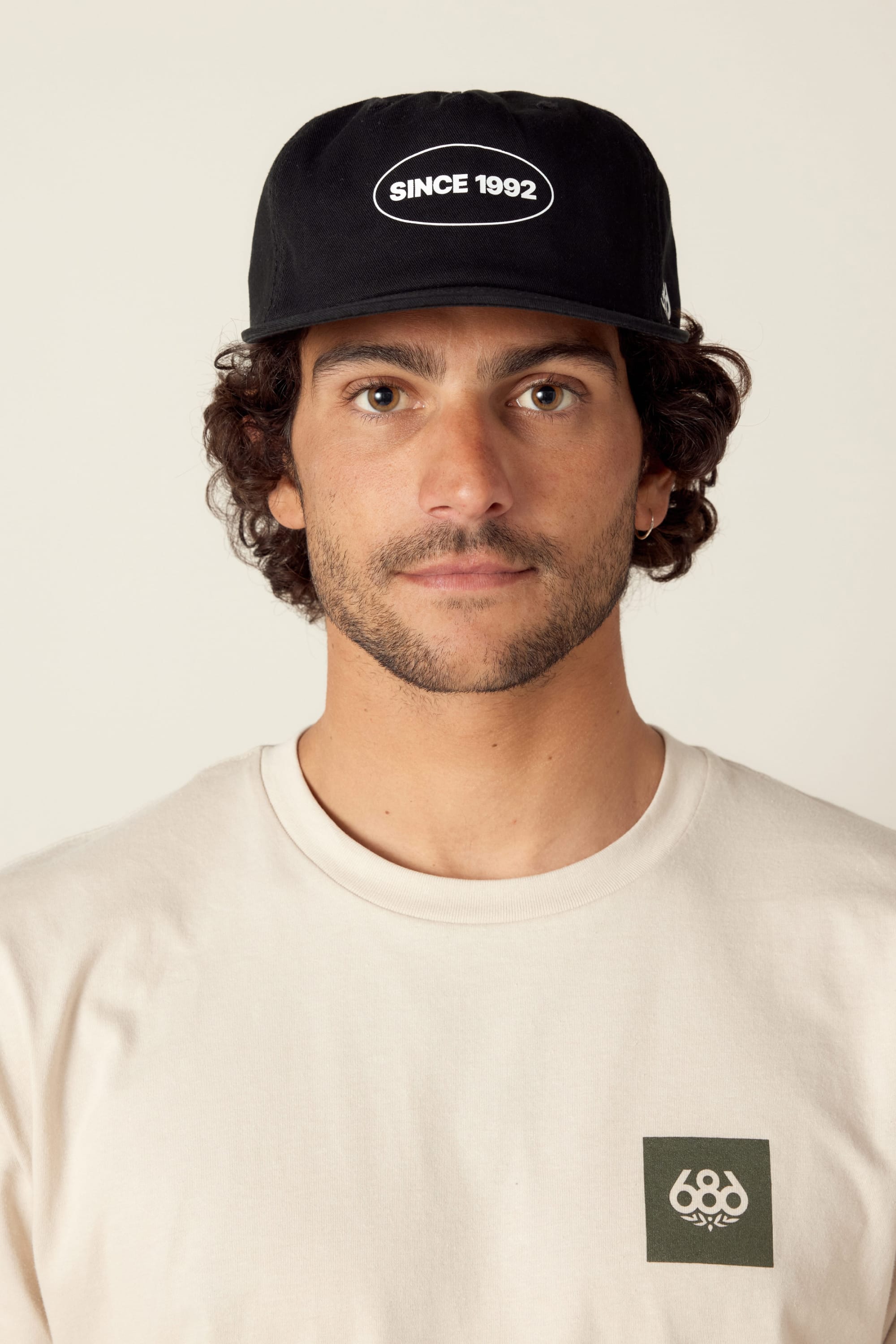 A man with curly brown hair and light facial hair wears the black 686 1992 Unstructured Hat and a beige T-shirt with a green logo, looking directly at the camera against a plain background.
