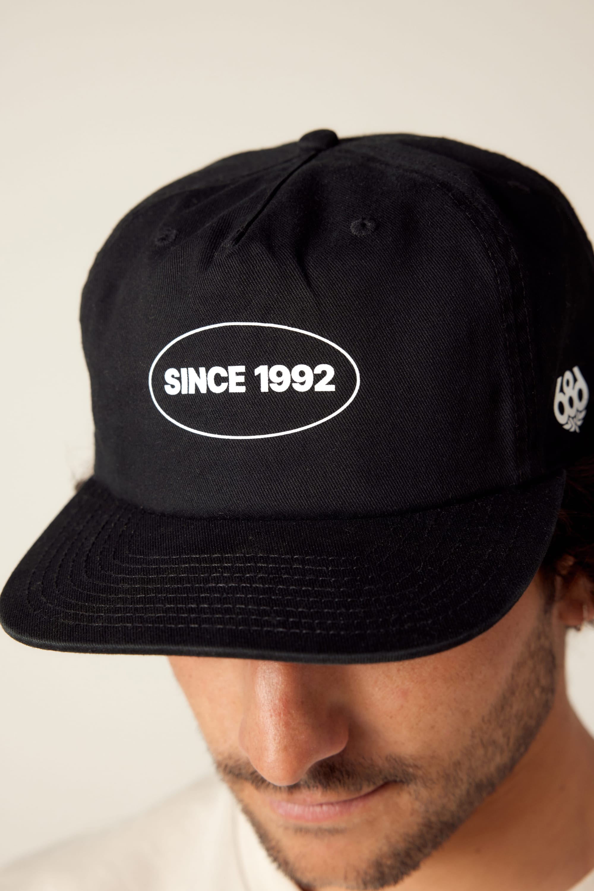 A person wearing the 686 1992 Unstructured Hat—a black men’s cotton hat by 686 with white “SINCE 1992.” text in an oval. Only the top of their face and the hat are visible.