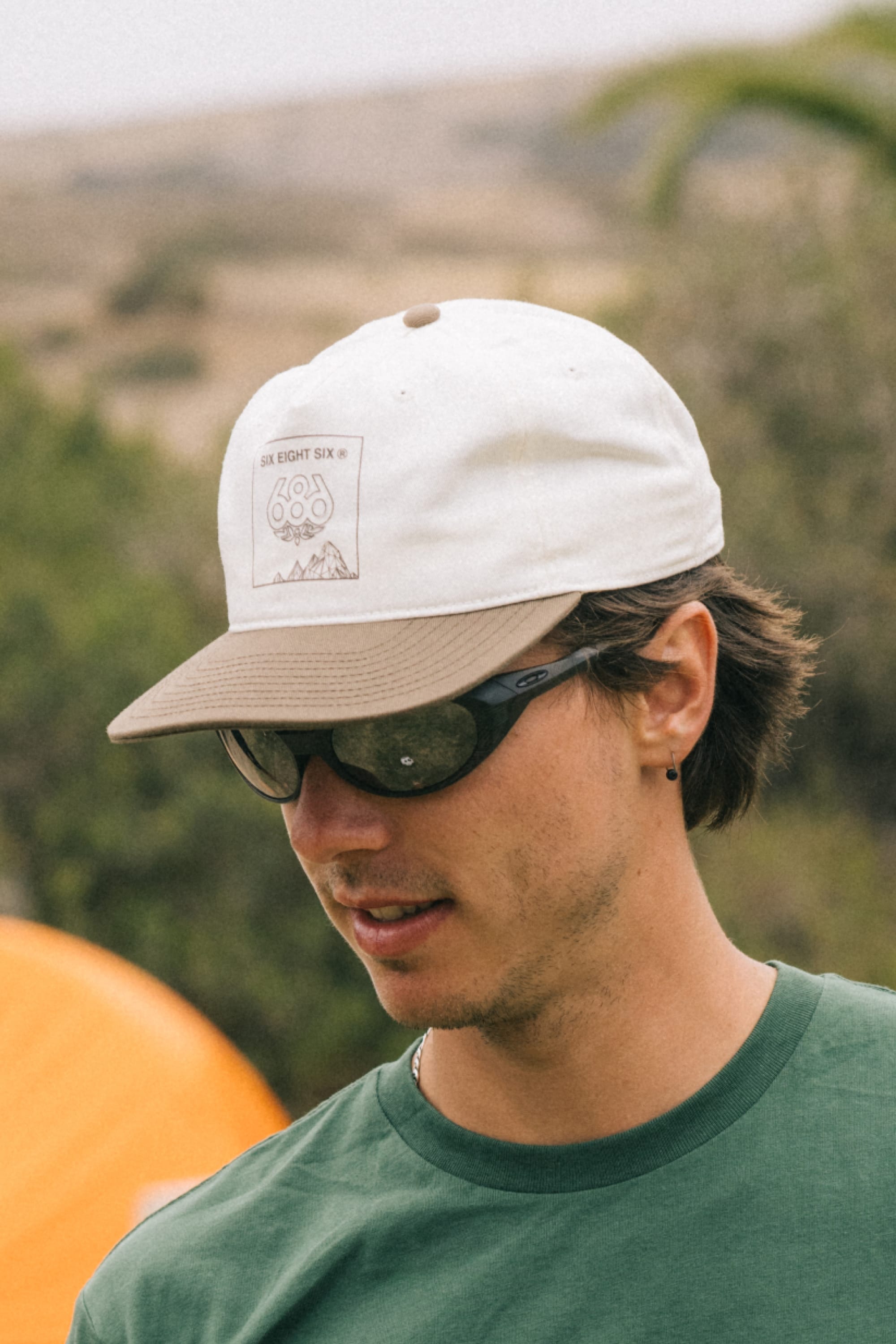 A young man stands outdoors in front of an orange tent, wearing a green shirt, dark sunglasses, and the 686 Cross-Cut Unstructured Hat by 686, with a blurred natural background.
