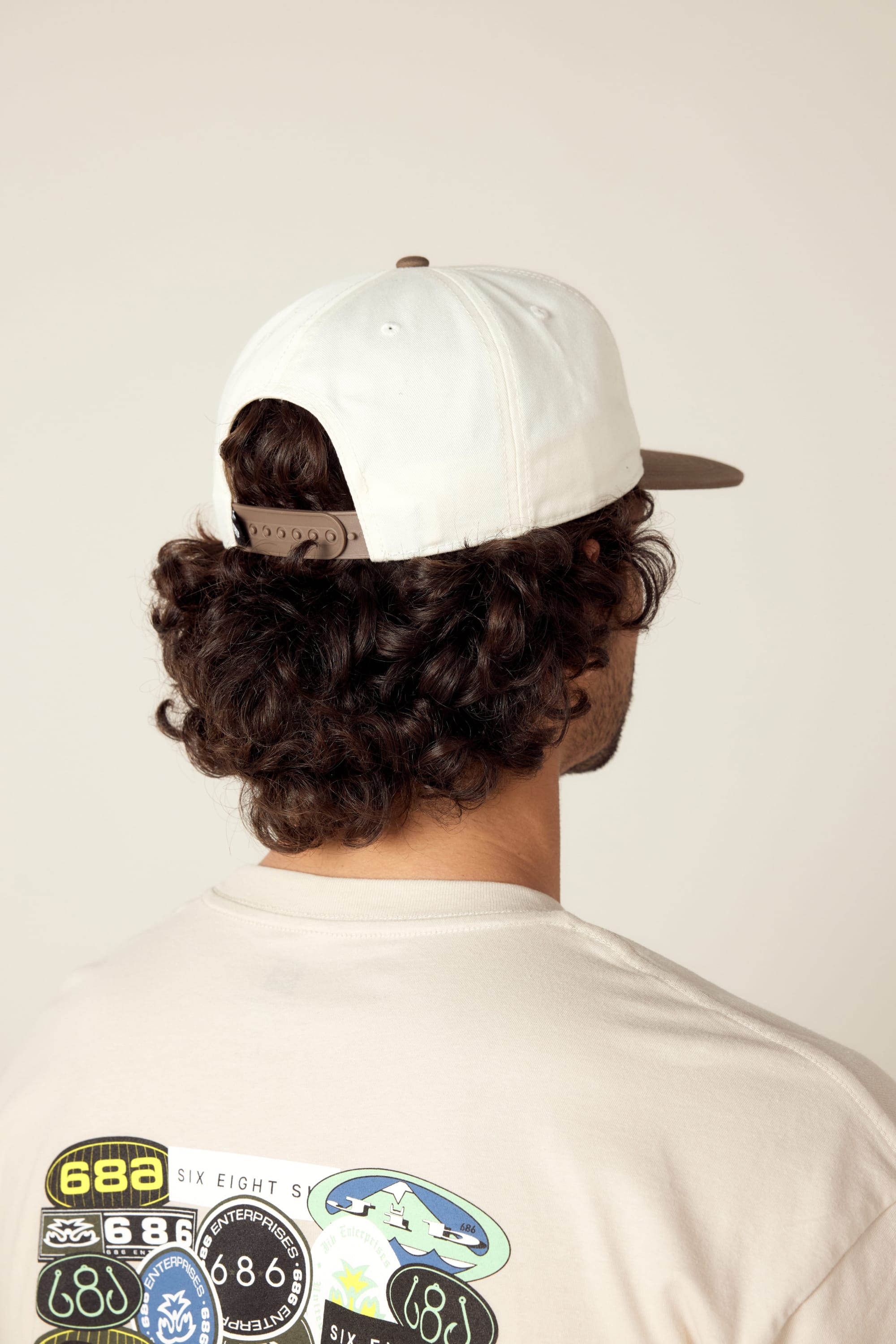 A person with curly hair is seen from behind wearing the 686 Cross Cut Unstructured Hat by 686, a white cotton hat, and a light shirt featuring multiple colorful patches on the back against a plain, neutral background.