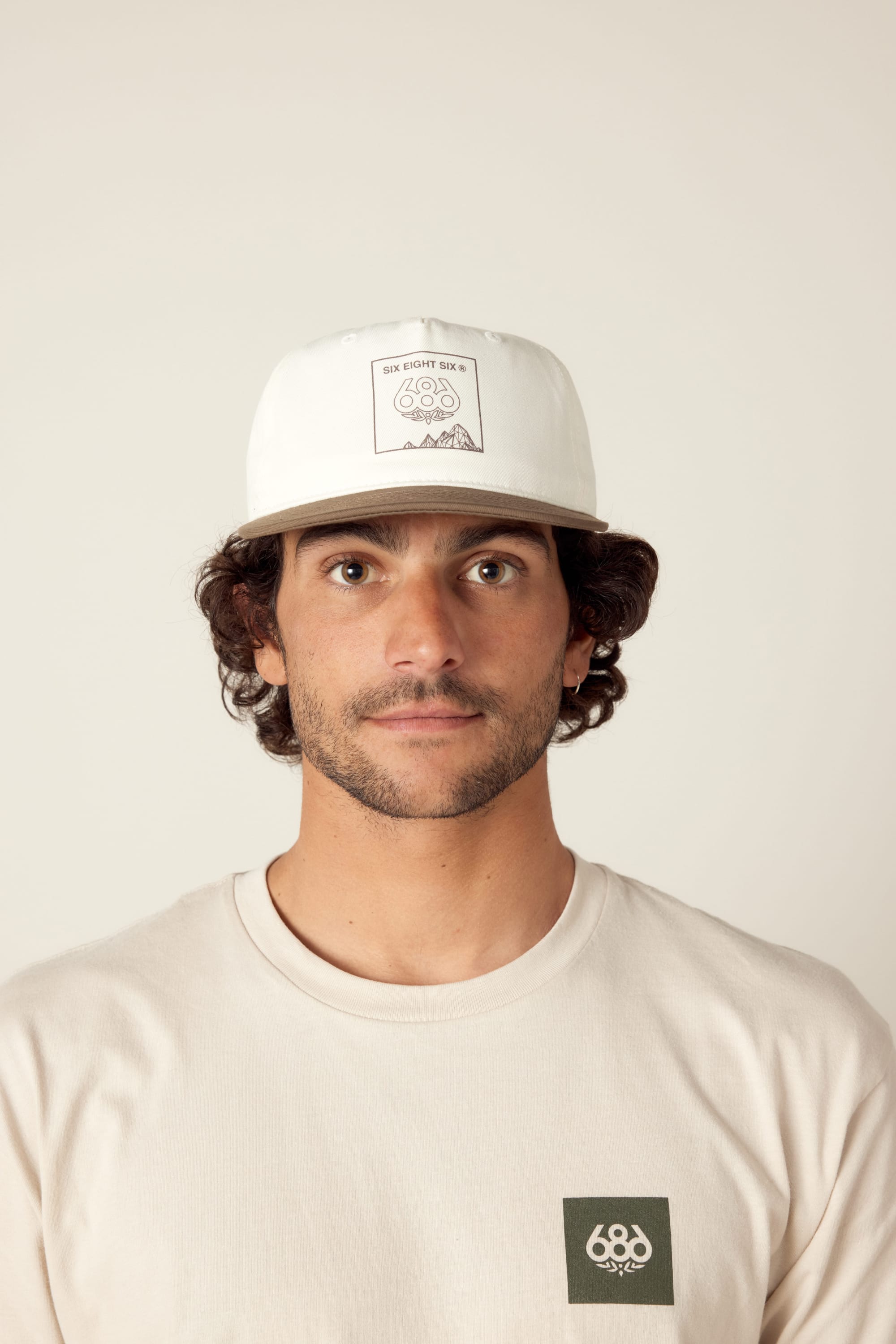 A man with wavy brown hair and light stubble faces the camera against a plain light background, wearing the 686 Cross Cut Unstructured Hat by 686, which is white with a graphic design, and a beige T-shirt with a small logo.