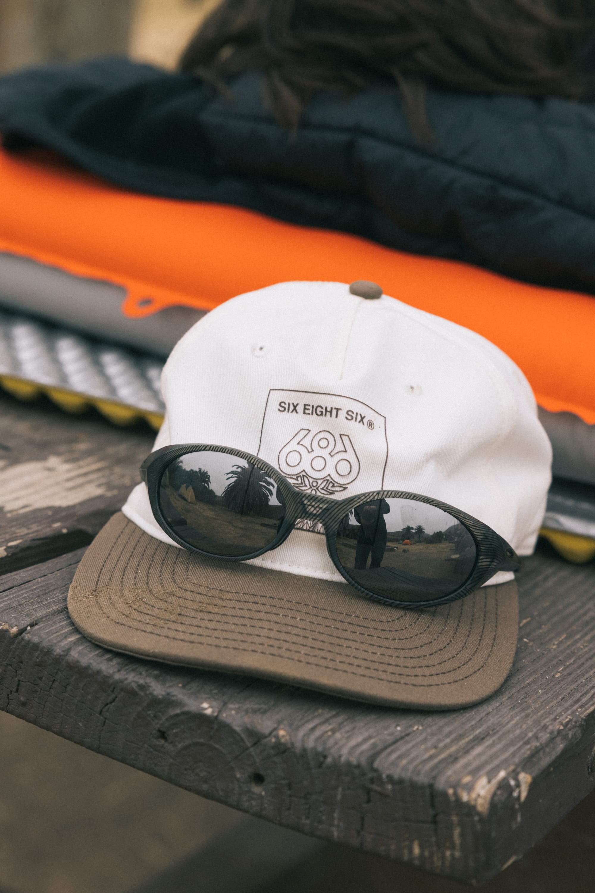 A 686 Cross-Cut Unstructured Hat in white and brown with a 686 patch sits on a wooden surface, black sunglasses across the brim. Folded clothes are stacked in the background.