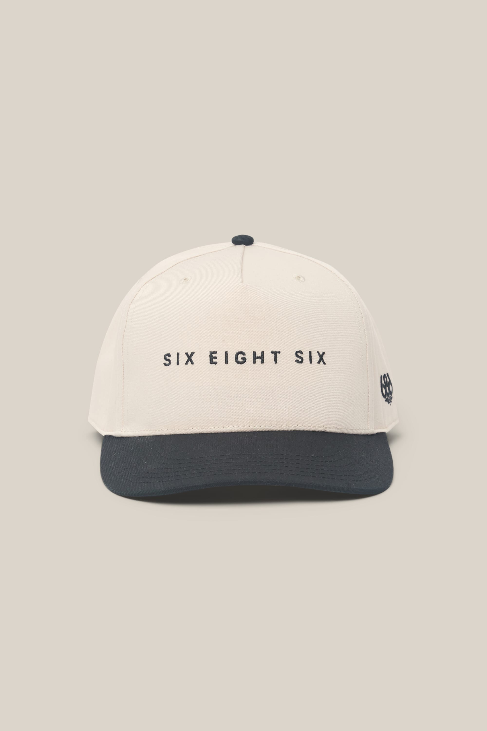 The 686 High-Pro Ses Two Tone Structured 5 Panel Hat by 686 is a men's white baseball cap made from 100% cotton, featuring a black brim, bold black SIX EIGHT SIX text on the front, and a small black logo on the left side on light beige.