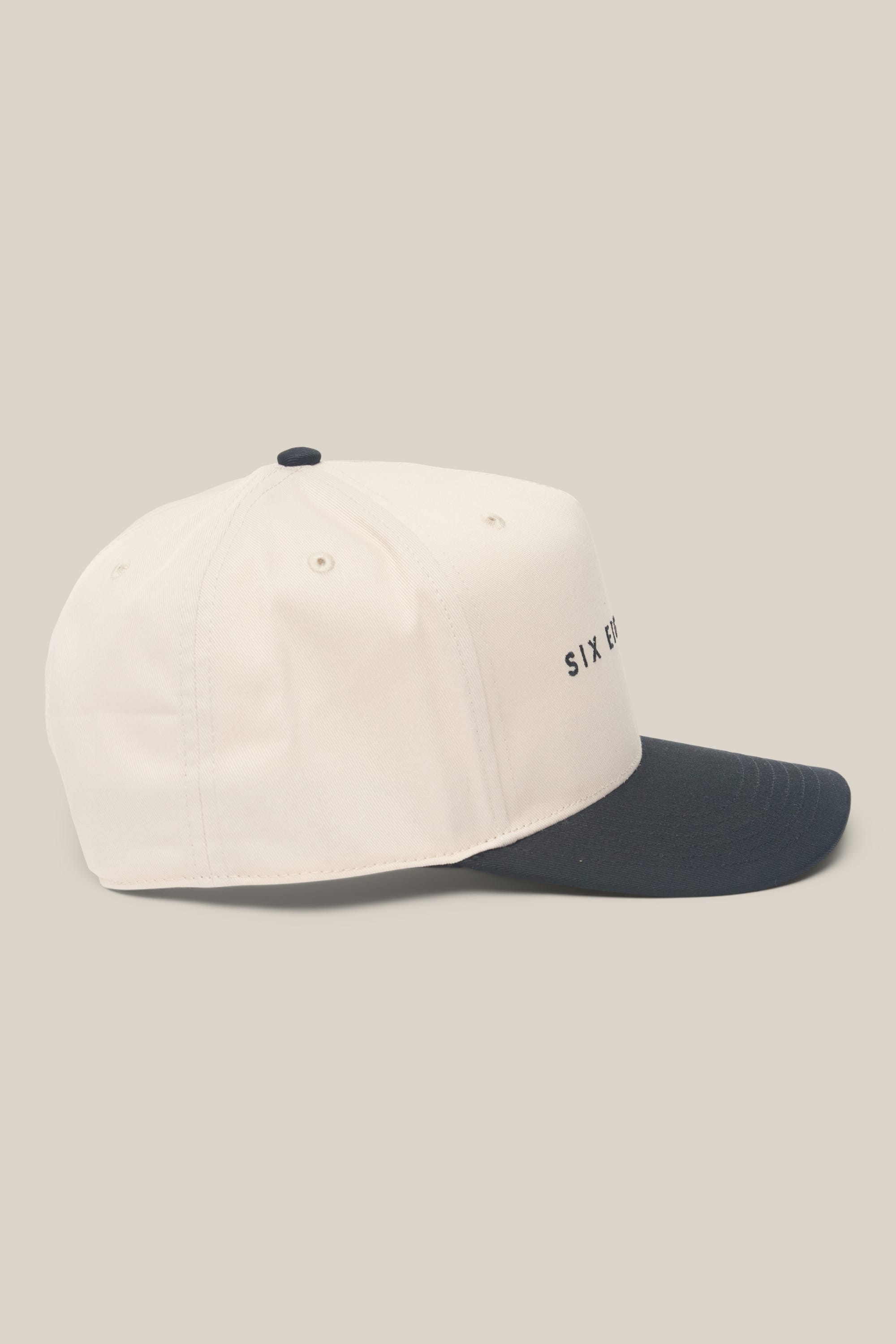 The 686 High-Pro Ses Two Tone Structured 5 Panel Hat by 686 is a men's white cotton baseball cap with a black curved brim, side ventilation eyelets, and partially visible SIX E text for a classic look.