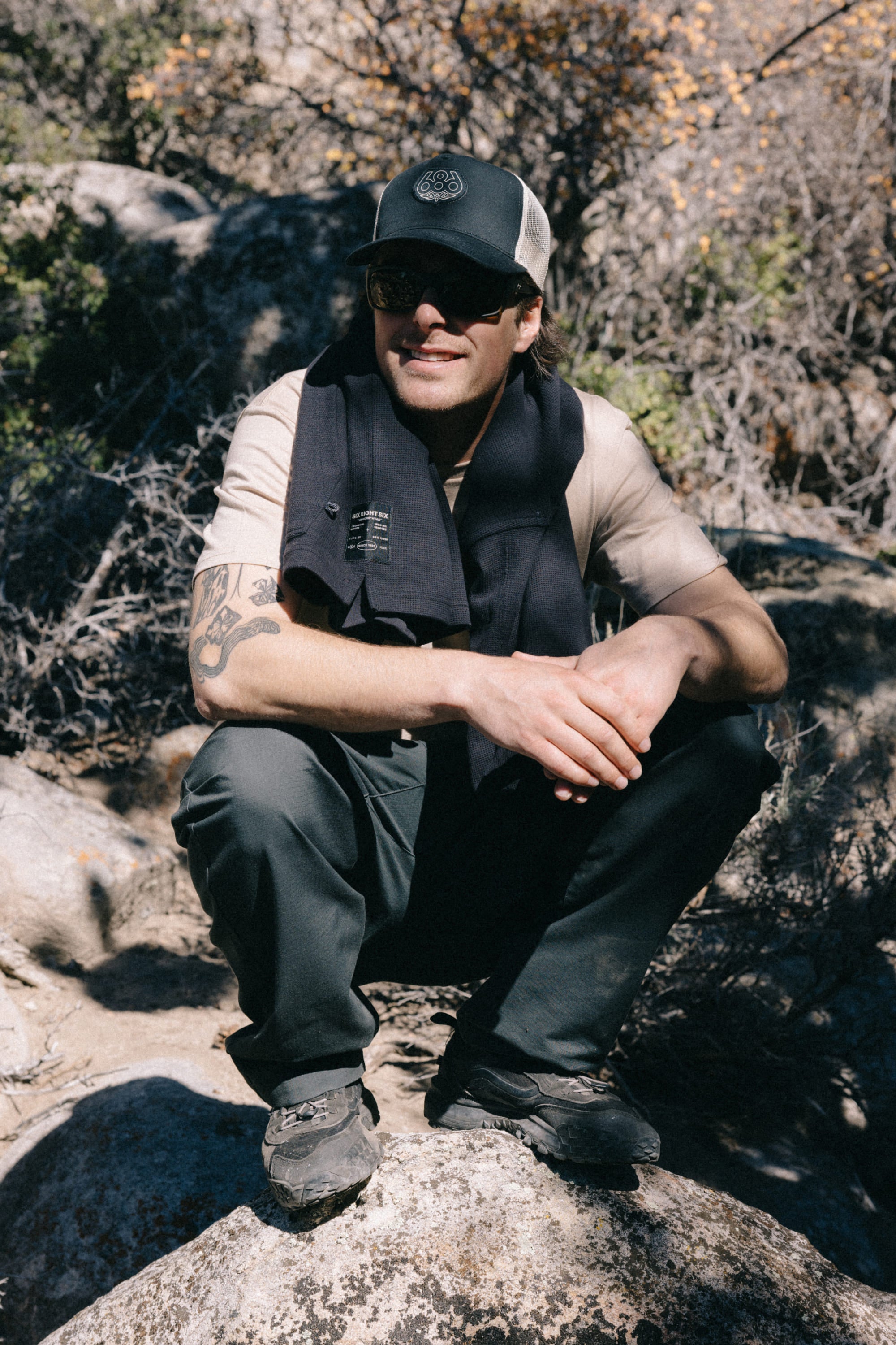 A person wearing the 686 Mid-Pro Wreath Unstructured 5 Panel Mesh Hat by 686, along with sunglasses and outdoor attire, squats on a rock in a sunlit, rocky landscape with sparse vegetation.