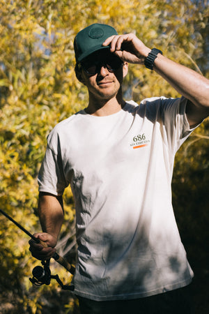 Wearing the 686 Mid-Pro Wreath Unstructured 5 Panel Mesh Hat, a person in sunglasses and a cap adjusts their hat while holding a fishing rod, standing outdoors in the sunlight with leafy trees in the background.