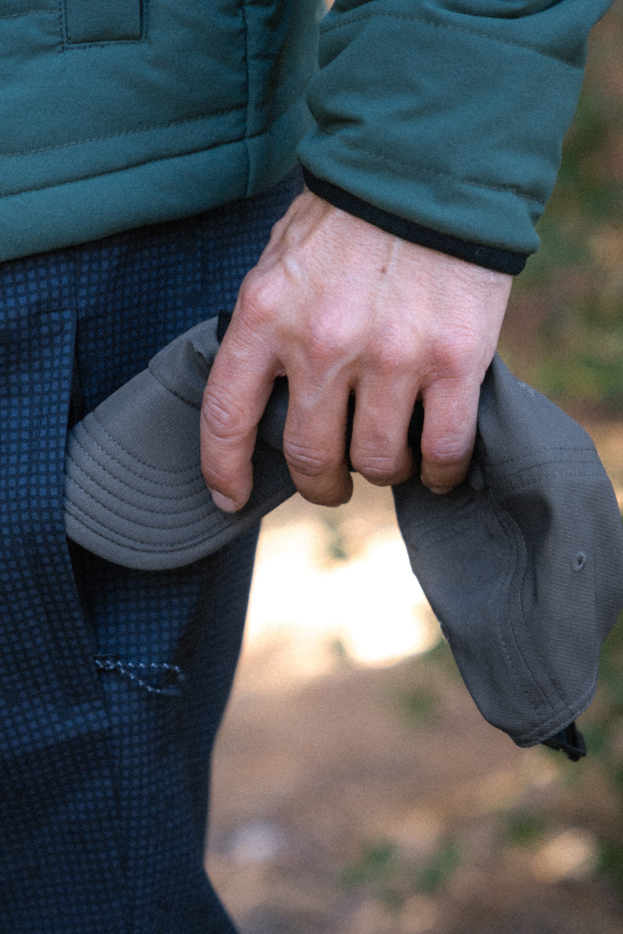 A person wearing a green jacket and blue checkered pants holds the 686 Low-Pro Adventure Unstructured 6 Panel Hat by the brim, their arm relaxed at their side.