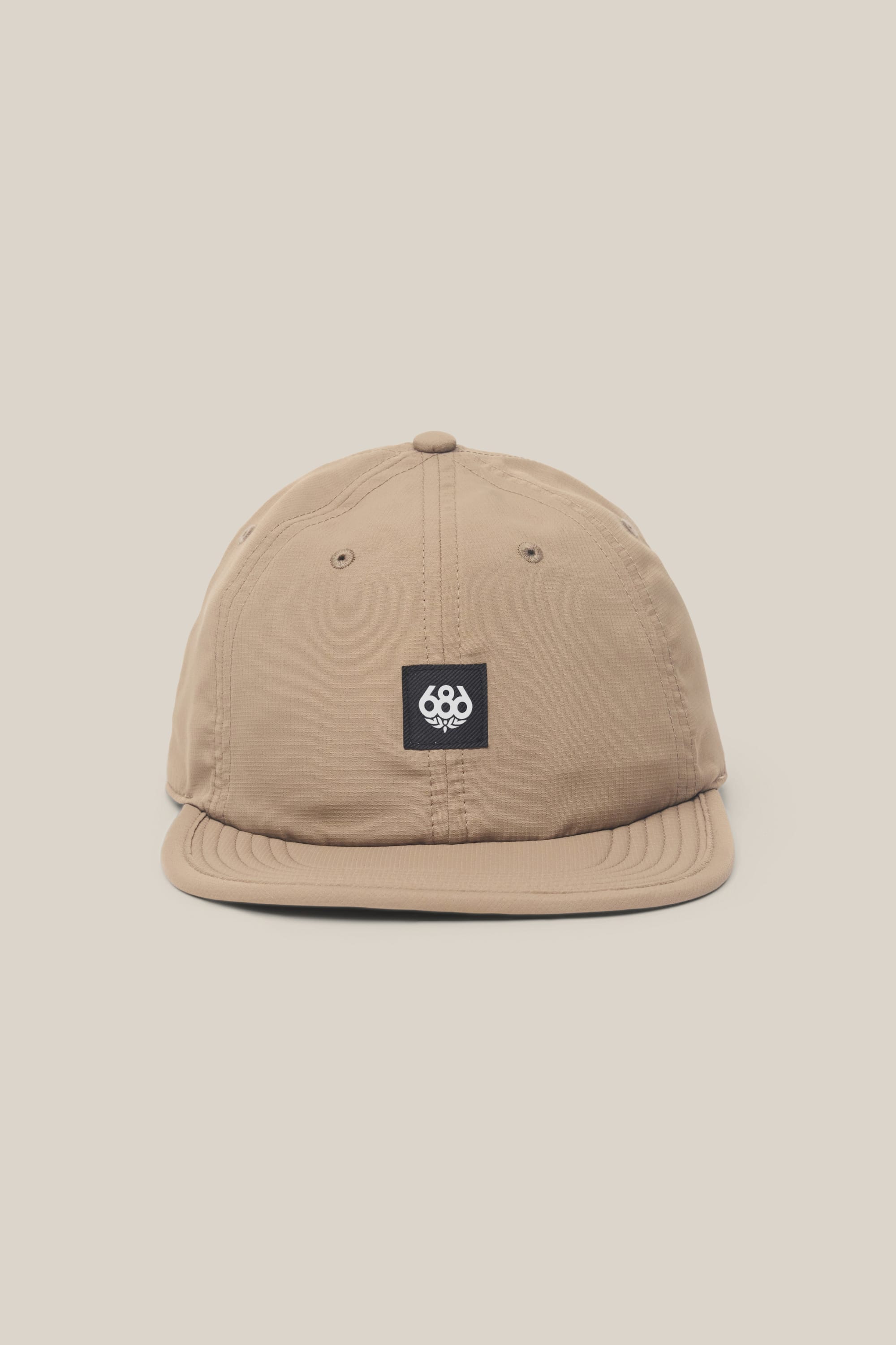 The 686 Low-Pro Adventure Unstructured 6 Panel Hat by 686 is a light brown men's polyester-cotton baseball cap with a flat brim and a black square patch with white embroidered logo on the front, shown against a plain beige background.