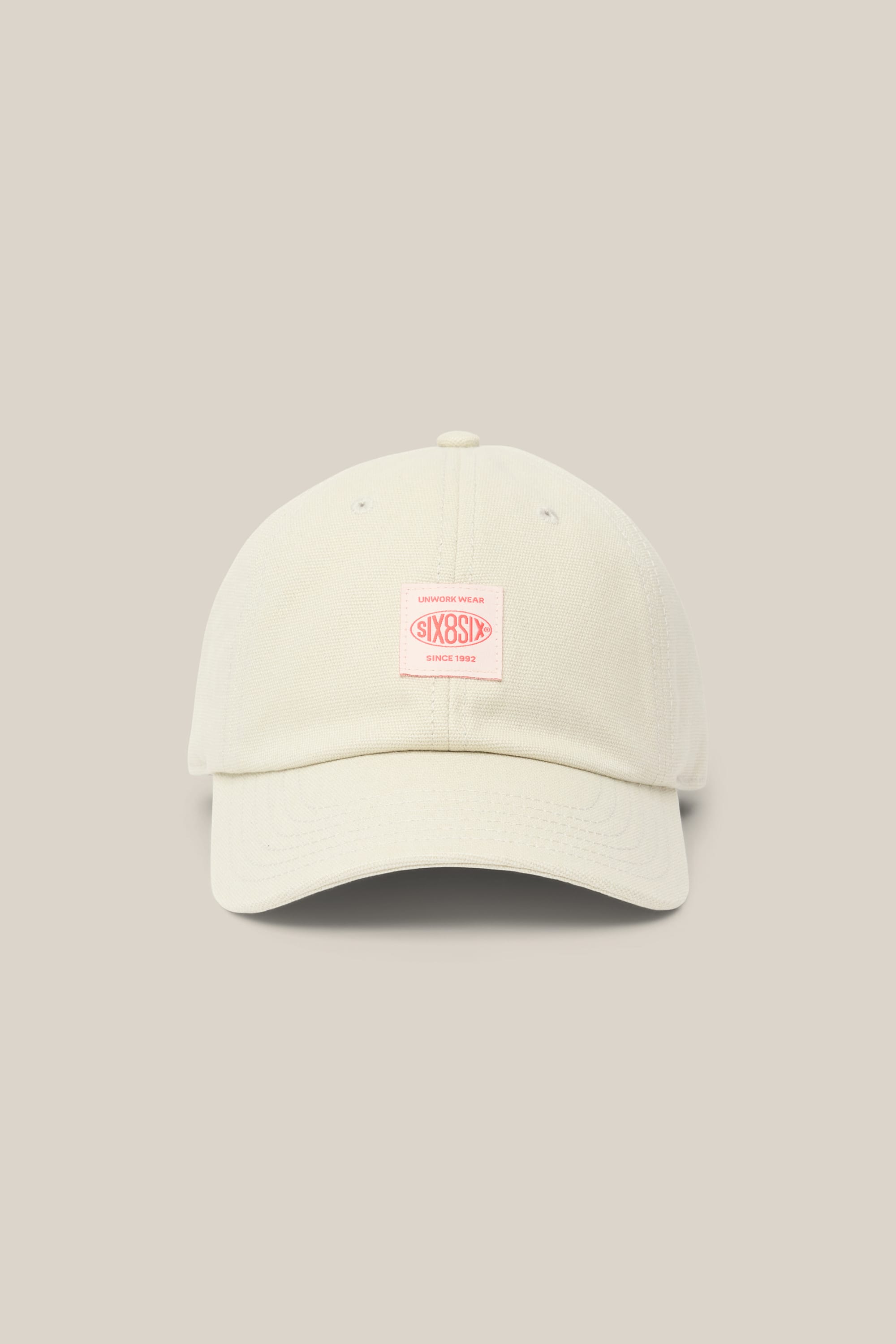 Introducing the 686 Low-Pro Unwork Unstructured 6 Panel Hat by 686—a men's baseball cap in a cream color, designed with a curved brim and adorned with a pink rectangular patch featuring text on the front. Made from a soft polyester cotton blend, this stylish hat is showcased against a neutral beige background.