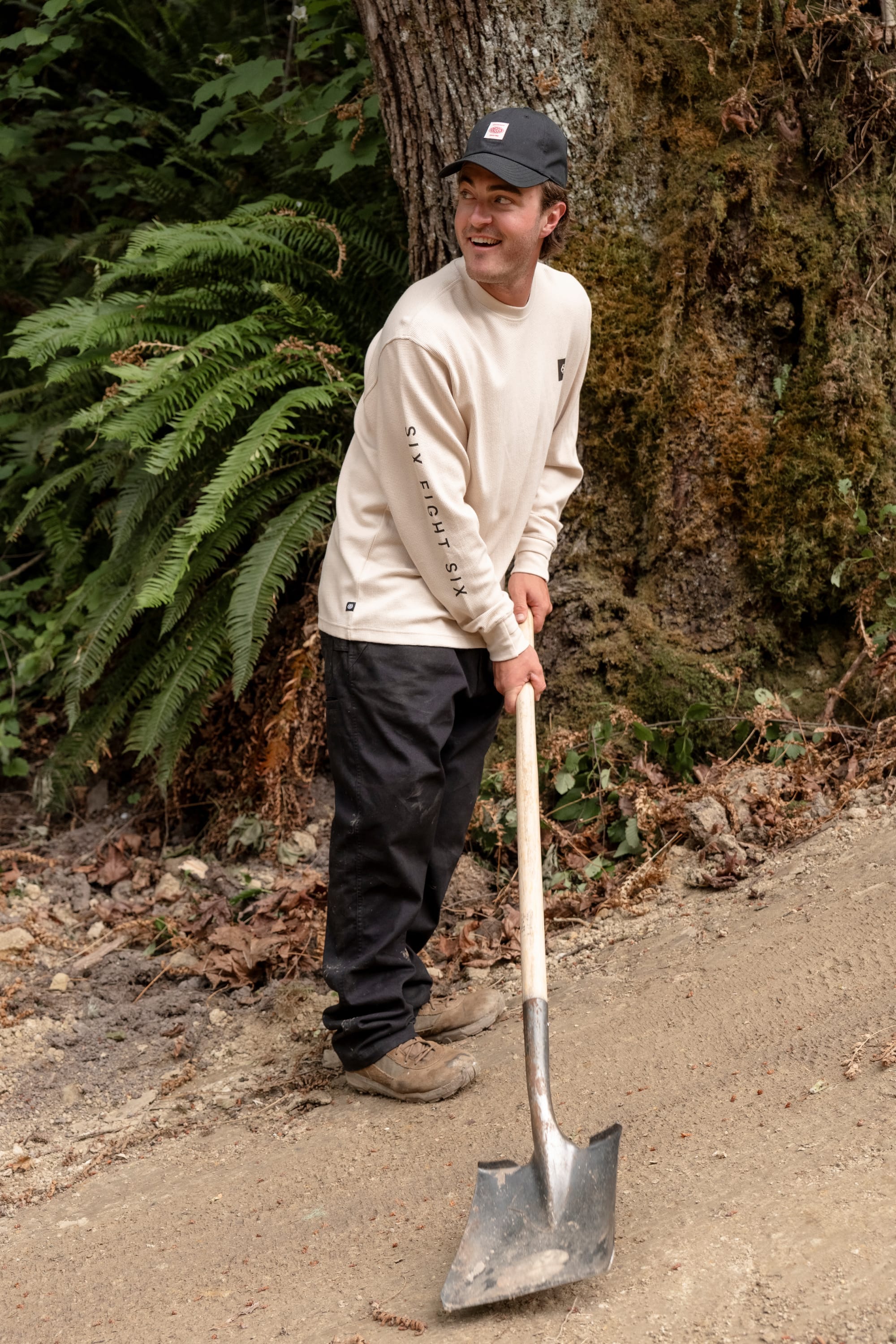 A person in a beige long-sleeve shirt, black pants, and a stylish 686 Low-Pro Unwork Unstructured 6 Panel Hat by 686 smiles while holding a shovel on a dirt path surrounded by trees and ferns.