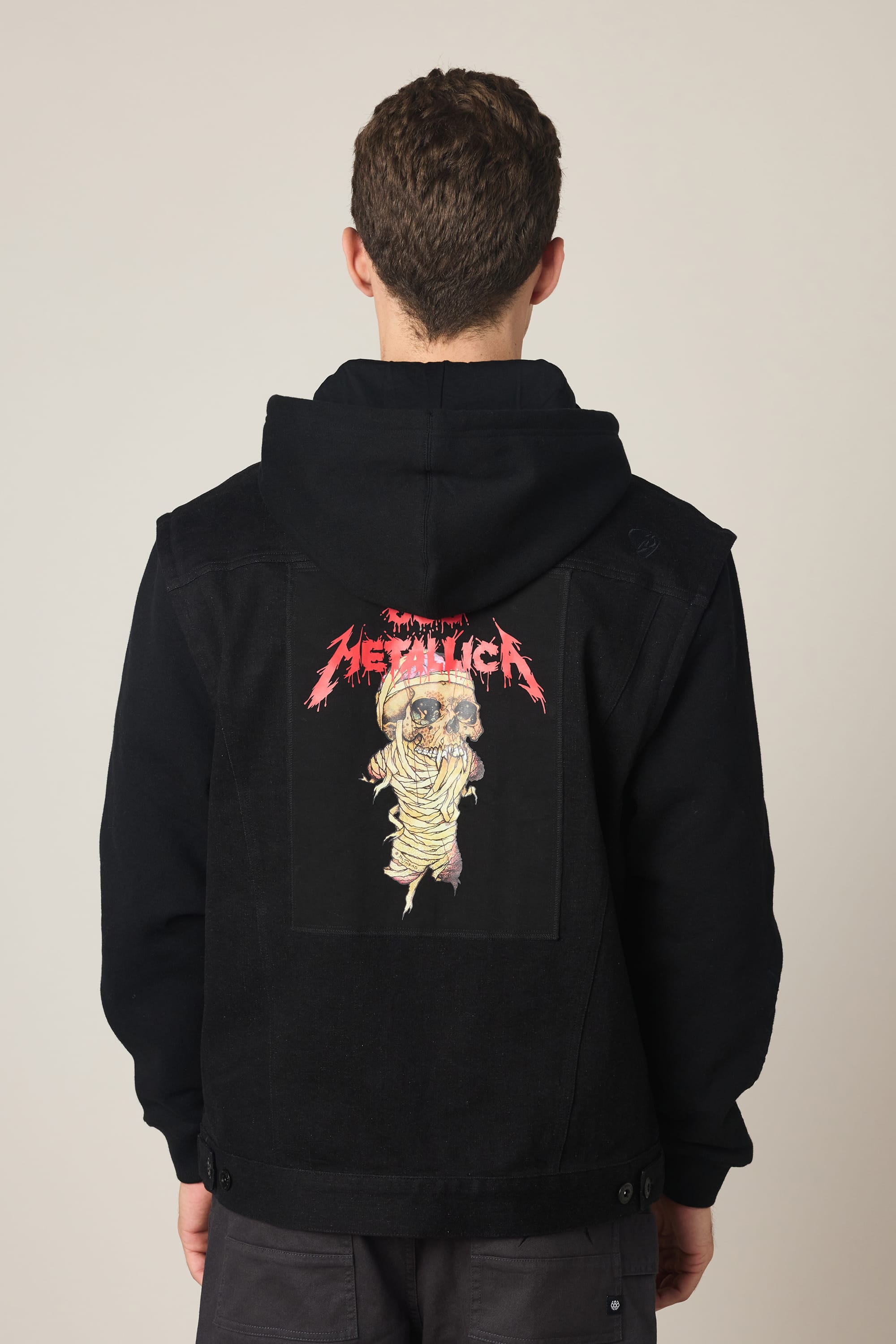A person is seen from behind wearing the 686 Men's Metalhead Trucker Jacket by 686, featuring a waterproof design and a striking Metallica graphic with bold red text and an illustrated mummy on the back.