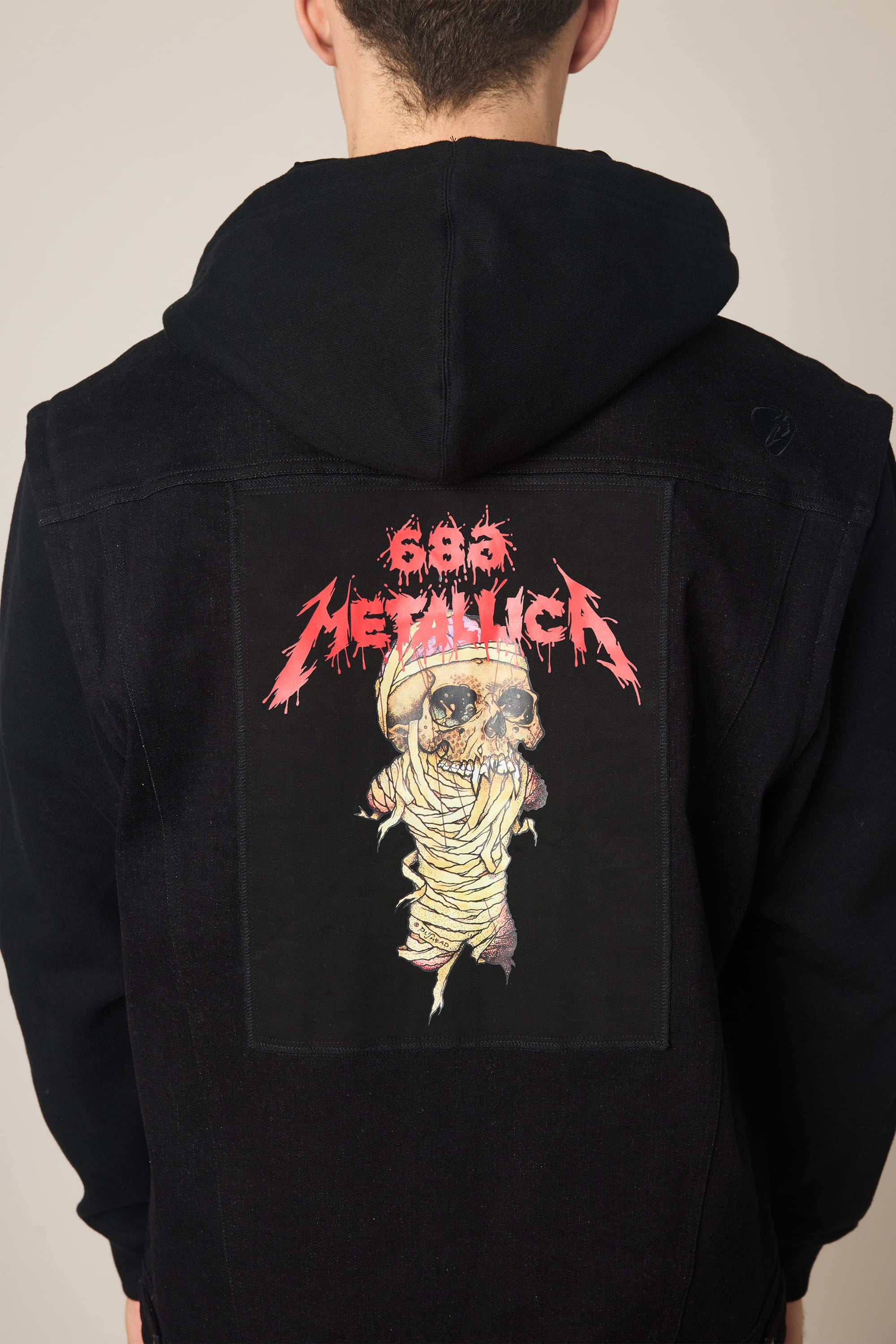 A person wearing the 686 Men's Metalhead Trucker Jacket, a black jacket by 686, featuring a large back patch with “Metallica” in red above a helmeted mummy skull.