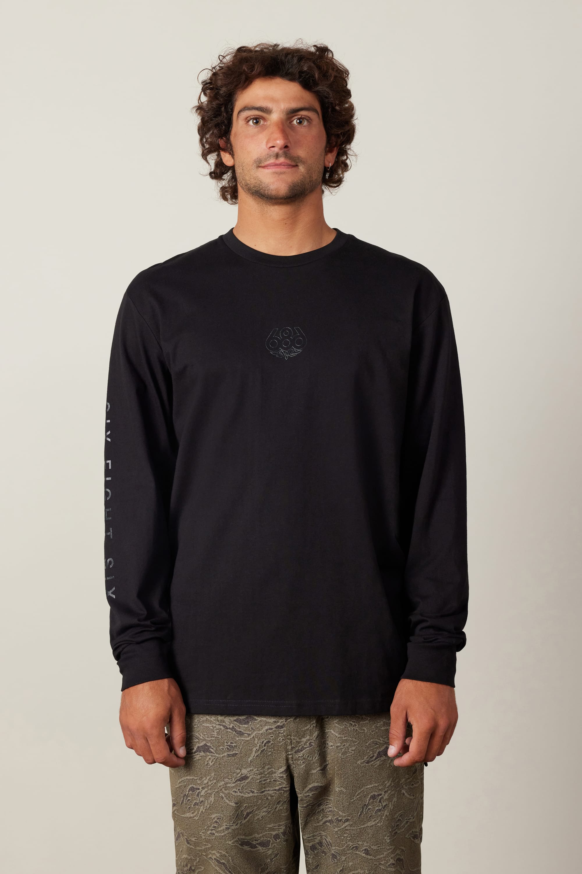 A man with curly hair wears the 686 High Def Authentic L/S Tee by 686, a black long-sleeve men's t-shirt with a subtle center logo and left sleeve print, paired with patterned olive-green pants against a plain background.