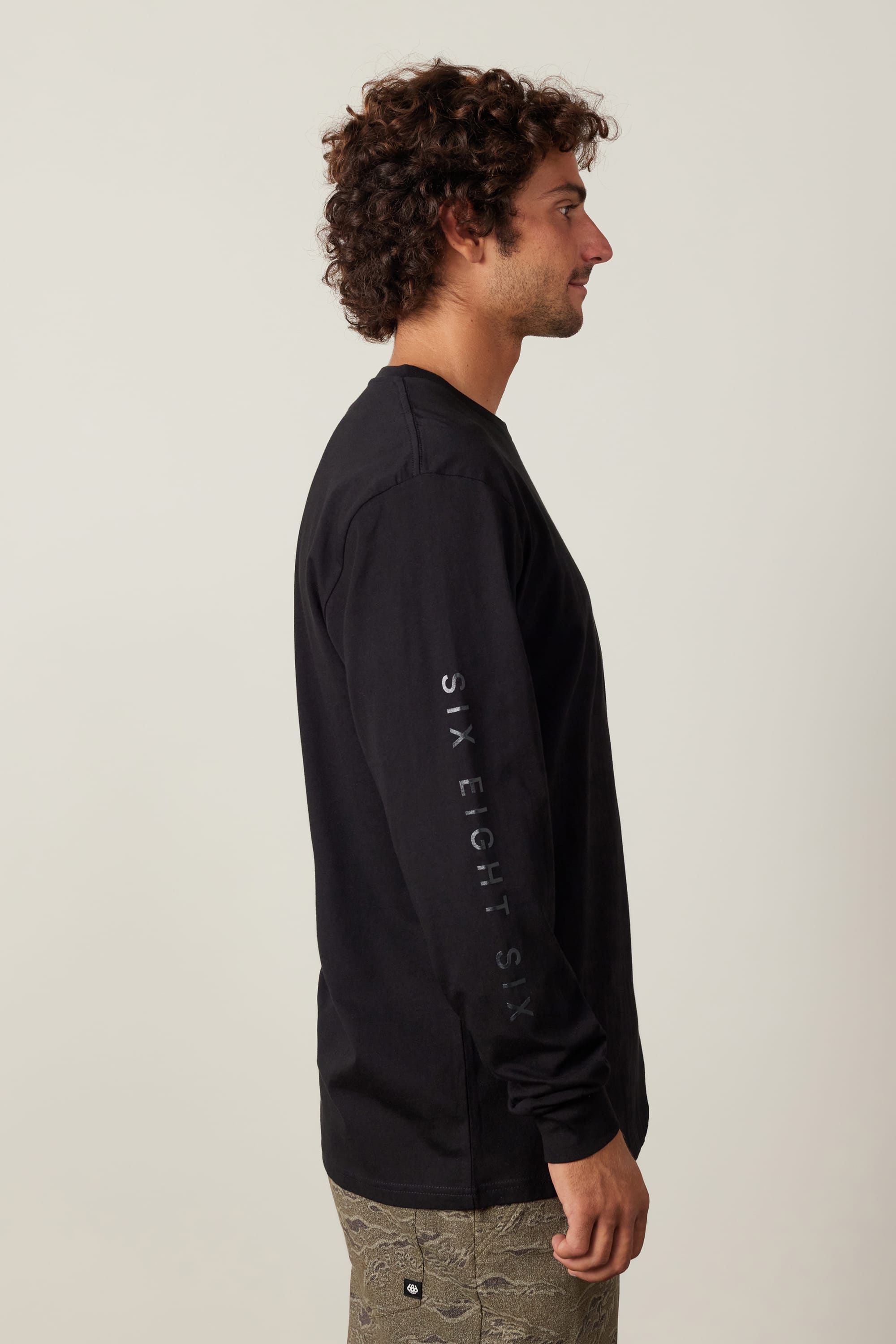 A man with curly hair stands in profile wearing the 686 High Def Authentic L/S Tee by 686, featuring "SIX EIGHT SIX" on the sleeve. His light patterned pants contrast the 100% cotton shirt, set against a plain light background.