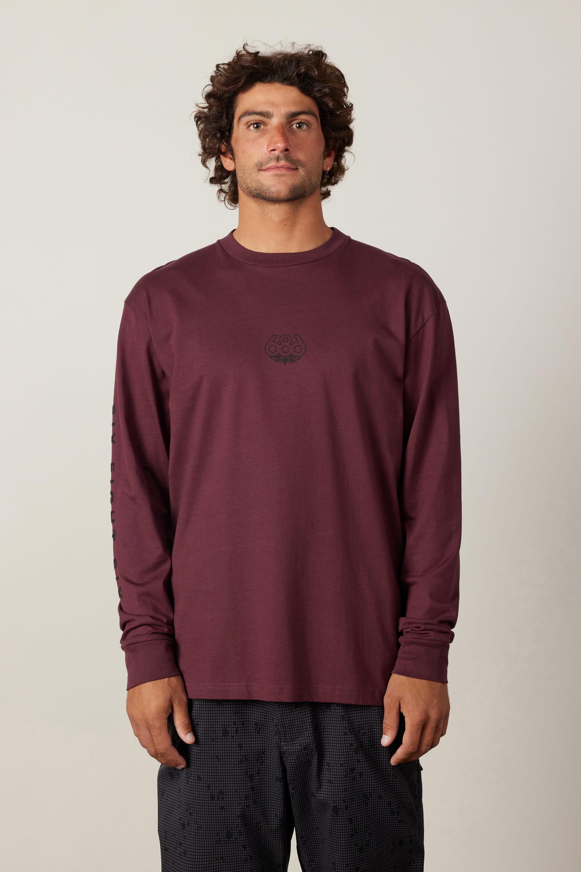 A man with curly hair wears the 686 High Def Authentic L/S Tee, a burgundy long-sleeve shirt with a small black chest logo by 686, paired with black patterned shorts, standing against a plain light background.