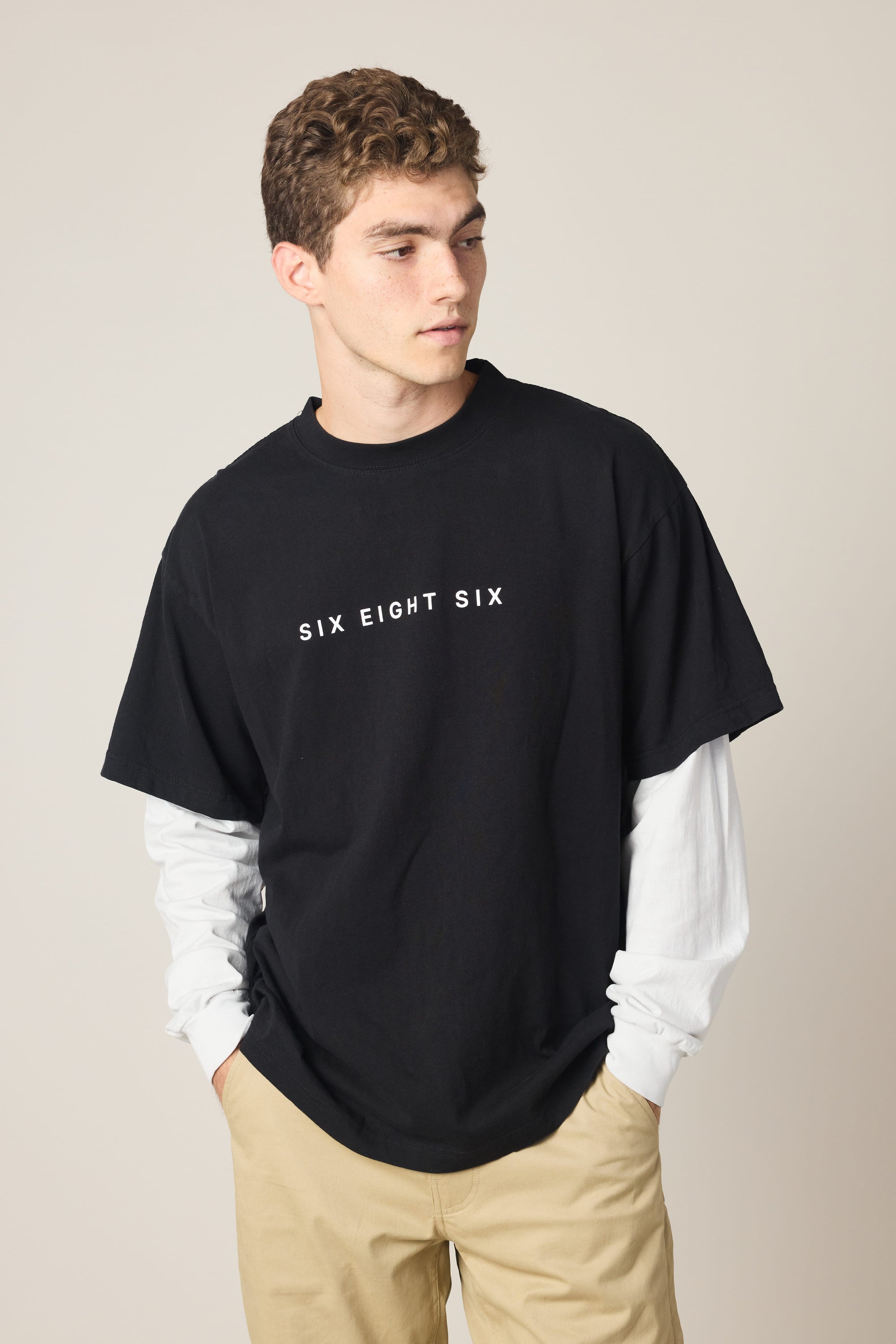 A young man with curly hair wears the 686 Wordmark Relaxed 2fer LS Tee by 686 over beige pants, glancing to the side with his hands in his pockets against a plain background.