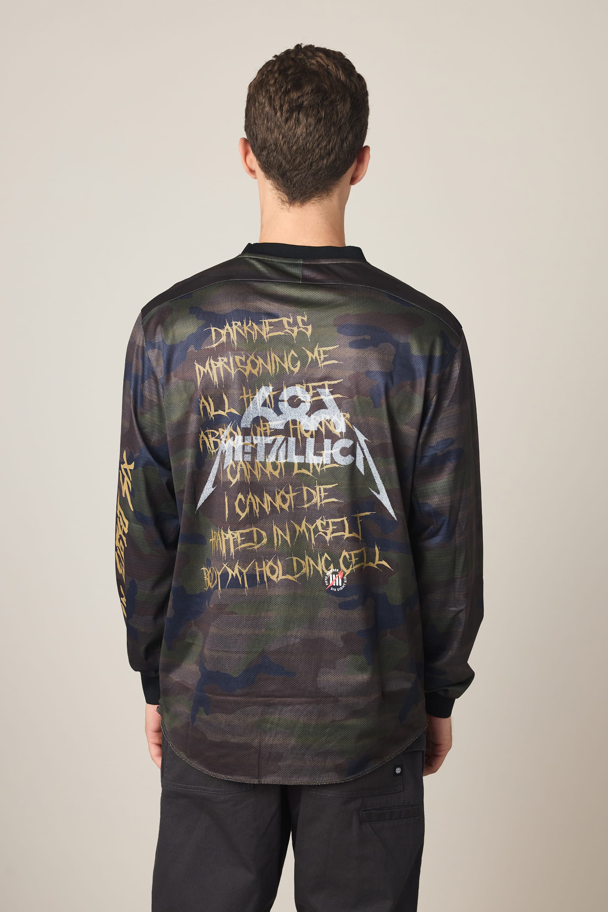 A person with short brown hair, seen from behind, wears the 686 Men's Metallica Turbo LS Tech Tee— a camo-patterned polyester shirt featuring Metallica’s logo, yellow graffiti-style song lyrics, and additional artwork and text on the back.