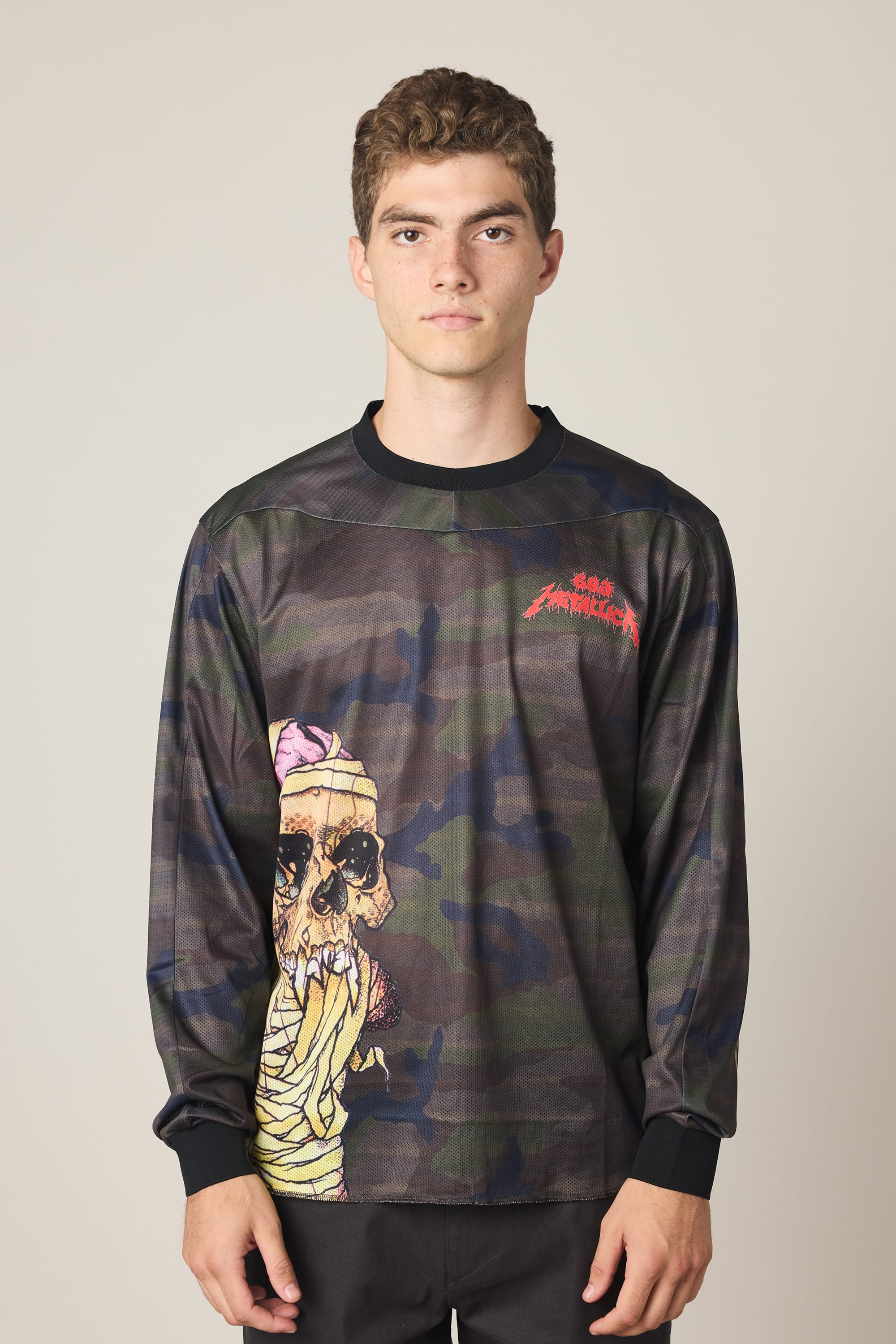 A young man with short curly hair models the 686 Men's Metallica Turbo LS Tech Tee, a long-sleeve, camouflage-style shirt with a bold skull graphic and red Santa Cruz text on the upper left chest, standing against a plain light background.
